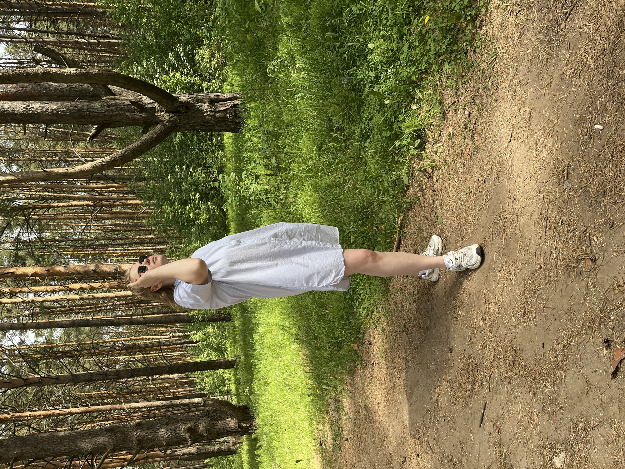 A young woman stands on a dirt path in a forest setting. She is wearing a short, light-colored dress and white sneakers. She is also wearing sunglasses. The background consists of tall pine trees and green foliage. The dirt path is covered with fallen pine needles. The location is Chaikovsky, Russia.