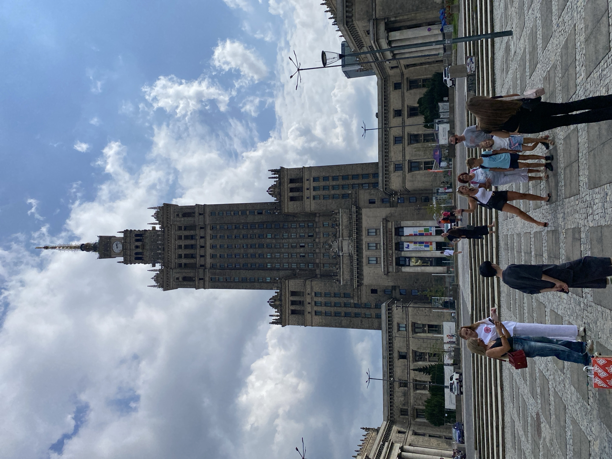 This image shows the Palace of Culture and Science, a prominent landmark in Warsaw, Poland. A group of people, consisting of adults and children, are standing in a plaza in front of the building. The building itself is a tall, Soviet-style skyscraper with a clock on its spire. The sky is partly cloudy. The people are casually dressed and appear to be tourists. Some are looking at their phones, while others are posing for photos. There are also cars and banners visible in the background. The plaza is paved with cobblestones and there are steps leading up to the building.