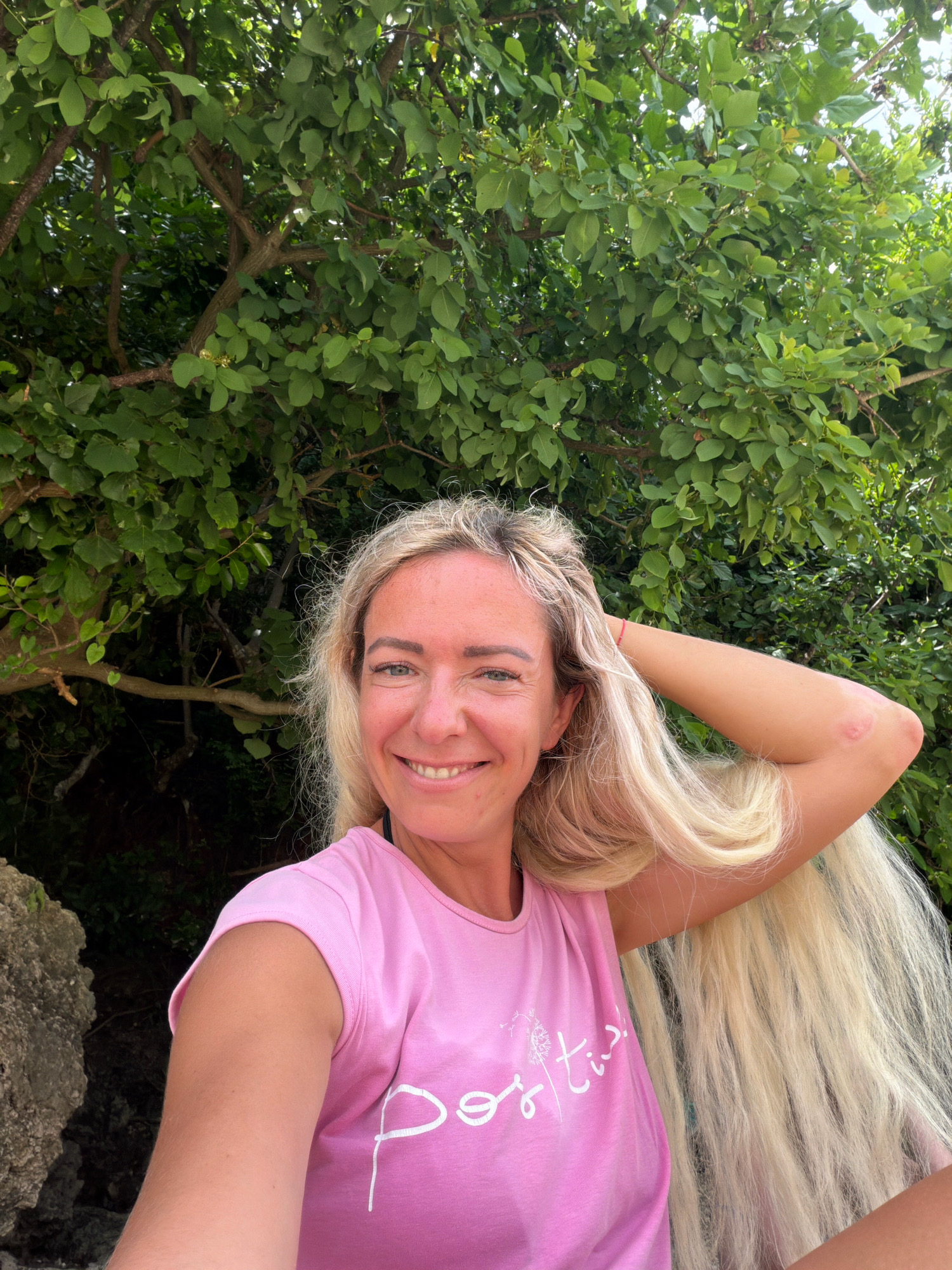 In Malay, Philippines, a blonde woman with long, flowing hair smiles as she takes a selfie in front of lush green foliage and a large rock. She is wearing a pink t-shirt with white text that reads "Positive". Her right arm is raised, with her hand resting behind her head. A red string bracelet is visible on her left wrist. The natural lighting suggests it is daytime.