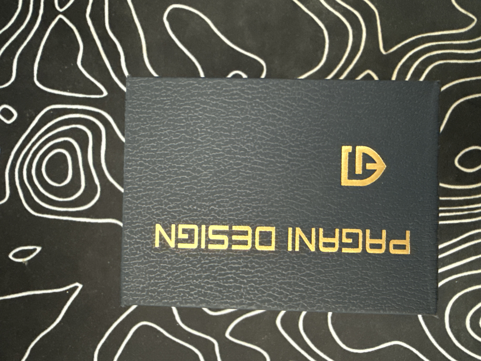 A dark blue textured box with gold lettering that reads "PAGANI DESIGN" is centered in the frame, placed on a black surface with a white topographic map-like pattern. The box is oriented vertically.