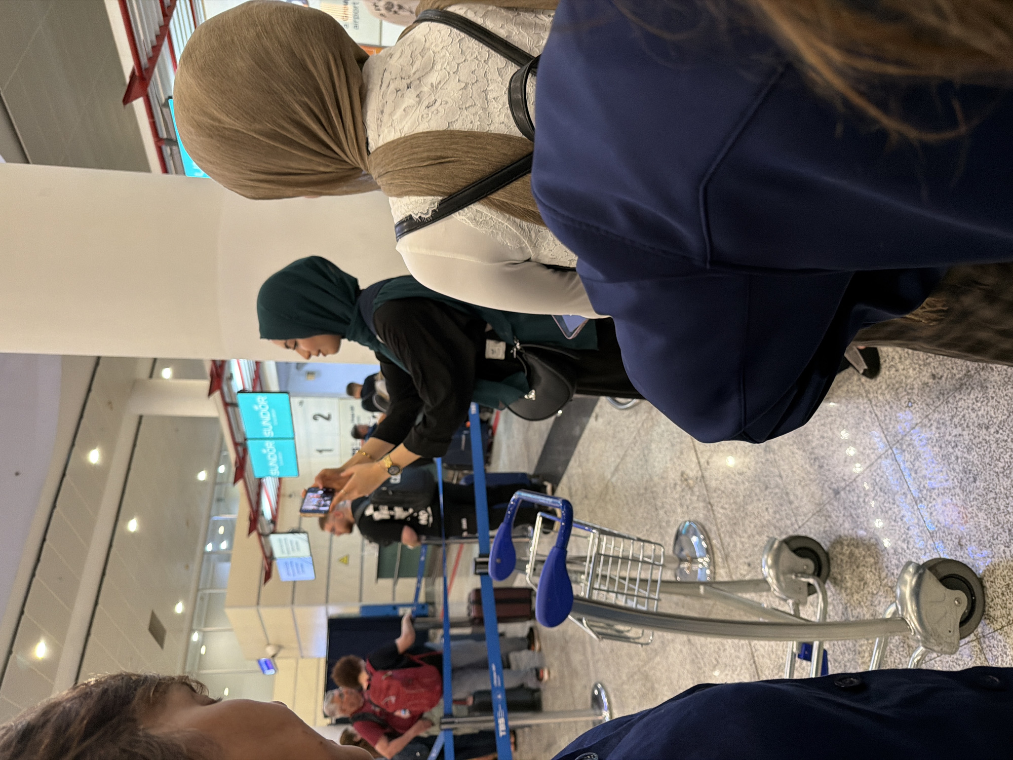 The image depicts a scene at Tbilisi International Airport, Georgia. In the foreground, two individuals are partially visible, one wearing a navy blue jacket. Behind them, several other people are standing in a queue, with luggage trolleys present. A woman in a green hijab is holding a mobile phone to take a picture. Another woman in a brown hijab is facing away from the camera. In the background, digital displays show flight information, including "SUNDOR SUNDOR," and numbers "1" and "2." The environment appears to be an airport terminal with tiled flooring and overhead lighting.