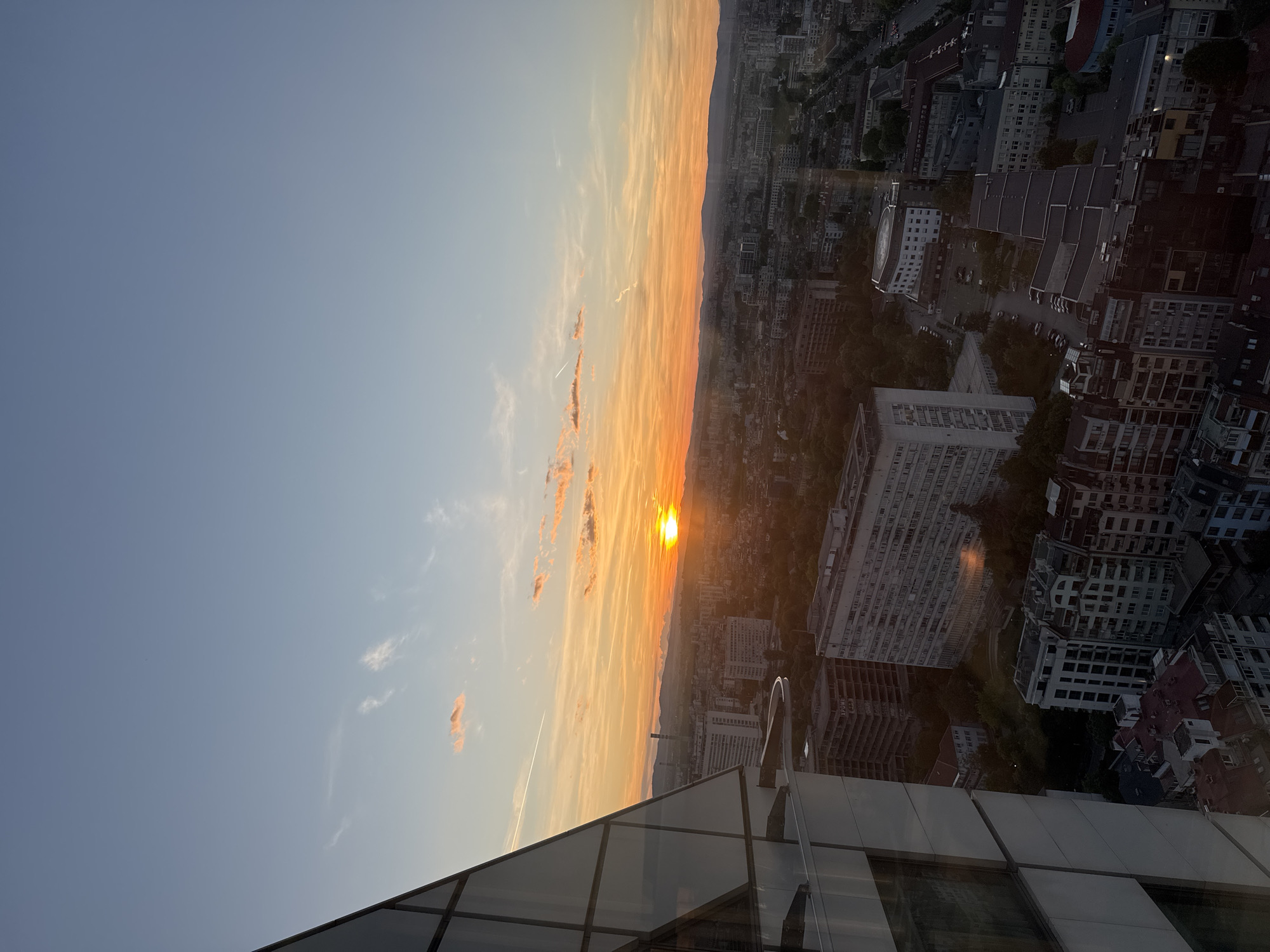 This media file presents a high-angle view of a sprawling urban landscape in Sofia, Bulgaria, captured during sunset. The image's dominant feature is the sky, transitioning from a clear, light blue at the top to vivid shades of orange and yellow near the horizon, where the sun is visible as an intensely bright, glowing orb. Several elongated, wispy clouds, illuminated by the sunset, stretch across the sky, along with a distinct airplane contrail.
Below the brightly lit horizon, a dense cityscape unfolds, revealing numerous buildings of varied architectural styles and heights. The urban area extends towards a dark, undulating mountain range or hills that are silhouetted against the colorful sky. Many buildings are multi-story, appearing as residential blocks or commercial structures, interspersed with areas of darker foliage, possibly parks or tree-lined streets. One prominent building in the mid-right appears to be under construction or unfinished, and faint Cyrillic text, possibly "ХРОМ," is discernible on its facade.
The immediate foreground features the glass facade and dark structural elements of a modern building, from which the photograph was taken. Reflections of the sky and distant cityscape are visible on these windows. The time of day is clearly sunset, indicated by the sun's low position and the warm, intense colors of the sky. The scene offers an expansive and peaceful view of the city as evening approaches.