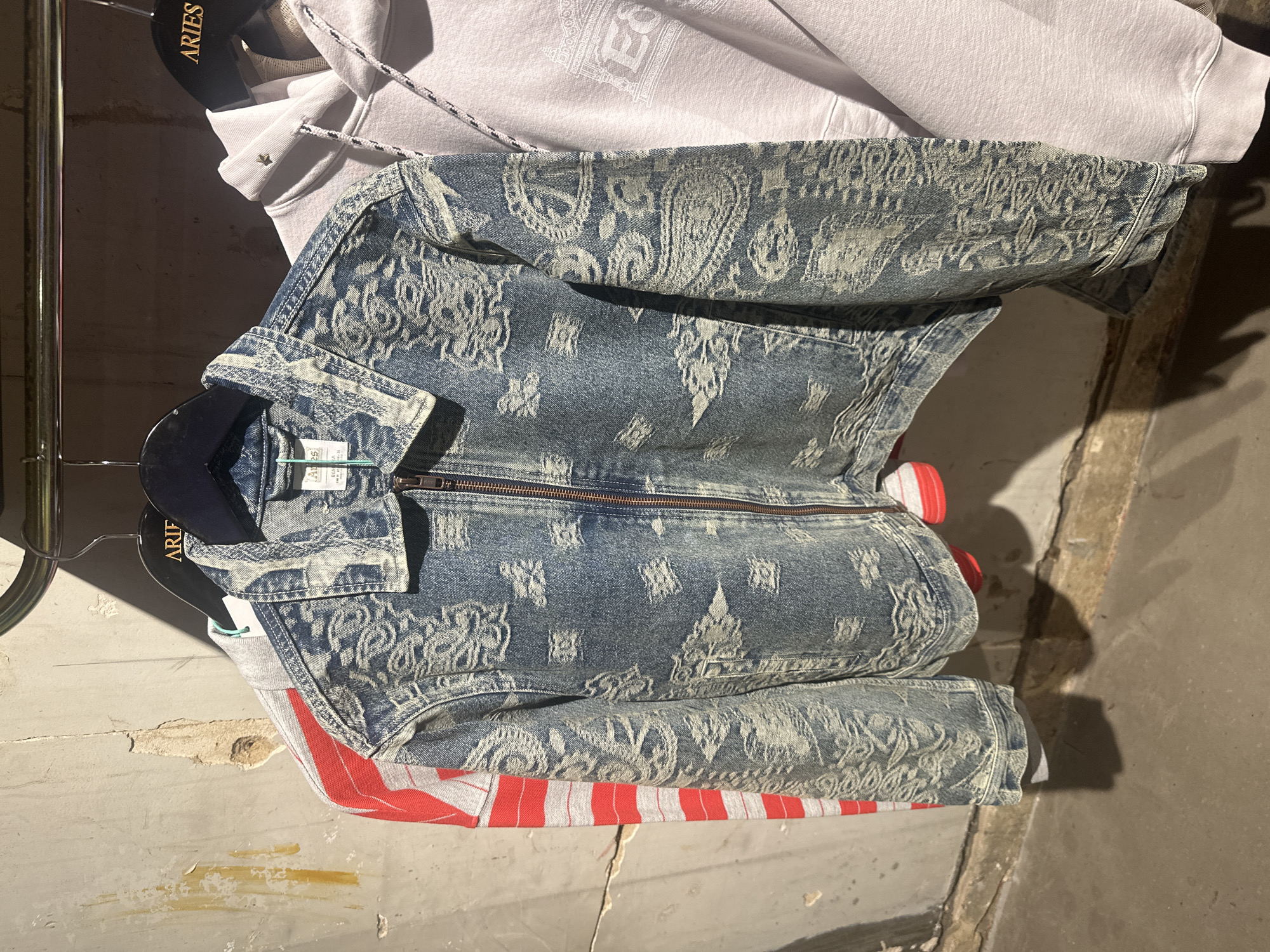 This close-up, vertical shot captures a display of clothing hanging on a metal rack, likely indoors. The scene features three main garments: a white hooded sweatshirt at the top with a patterned texture and rope drawstring, a blue denim-style jacket in the middle, and a red and white striped garment peeking out from the bottom.
The white hoodie has a small gold star detail near the drawstring and a faded, intricate pattern visible on its surface, which includes what looks like the letters "ES" or "E3" within a geometric design. The prominent blue jacket is made of a distressed, washed-out denim material, featuring an embossed or printed pattern consisting of paisleys and various geometric shapes in lighter blue and white tones. It has a visible zipper down the front and a classic collar. A small white label with some text, possibly size or brand information, is attached to the collar area of the blue jacket. The bottom garment is predominantly red with white horizontal stripes.
The clothing hangs from dark hangers, one of which clearly displays the brand name "ARIES" in white text. The background consists of a textured, distressed wall with visible cracks and worn areas, indicative of an older or industrial setting. The floor beneath is concrete. Strong lighting from the upper left creates distinct shadows on the wall and floor. No people are visible, and there is no direct activity taking place beyond the static display of garments. The overall impression is a retail or studio environment.