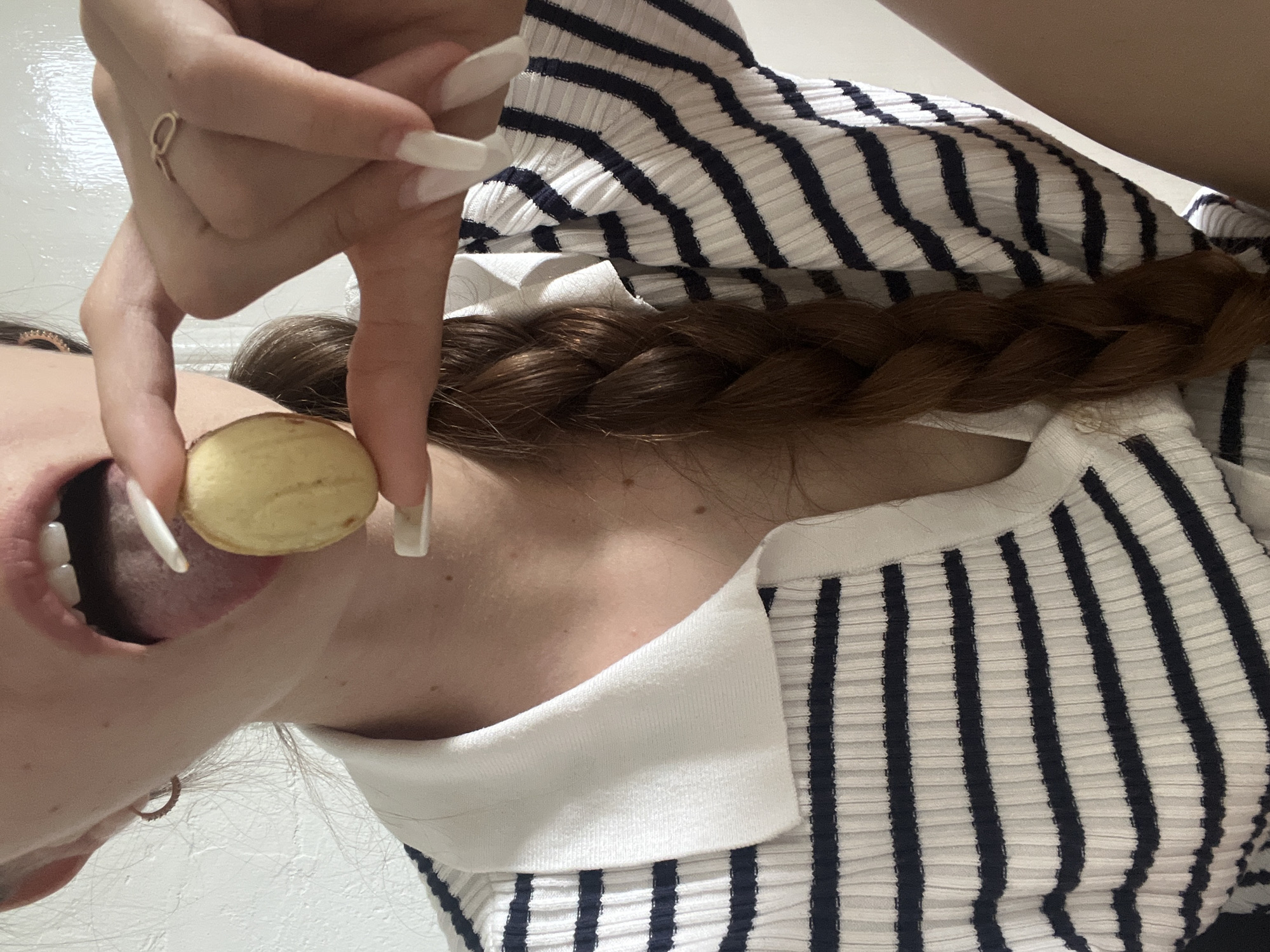 A close-up shot captures a person holding a small, light-colored, round object near their open mouth, with their tongue extending towards it. The person has long, light-colored fingernails and a braided brown hairstyle. They are wearing a white and navy striped collared shirt. The background is a plain, white wall.