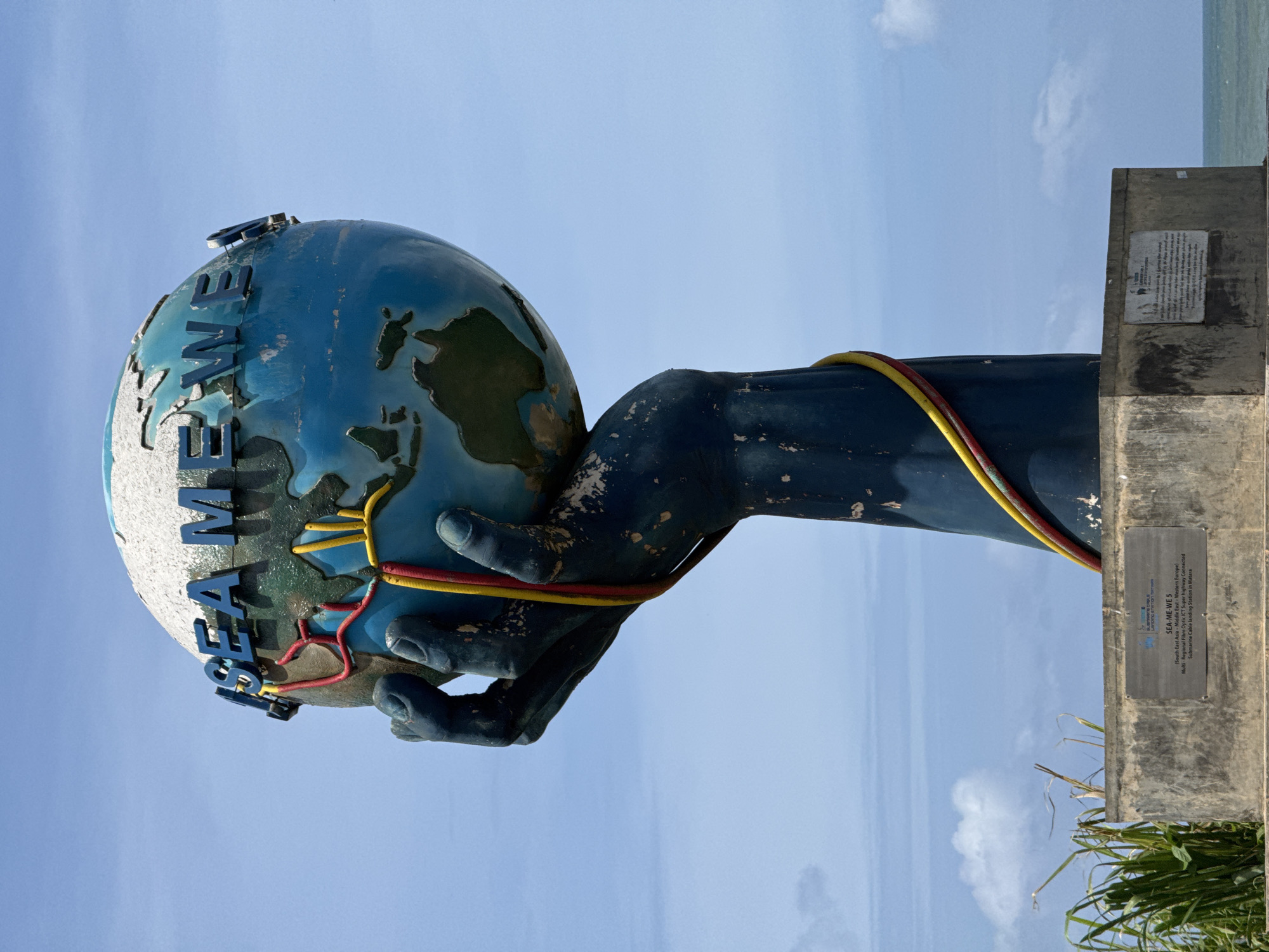 This is a full-shot, eye-level, outdoor photograph of a large monument in Dondra, Sri Lanka. The monument, which stands on a concrete base, depicts a dark blue, open hand holding up a globe of Earth. The globe has the words "SEA ME WE 5" written in blue lettering around its circumference. Red and yellow cables wrap around the hand and the globe, connecting to what appears to be a representation of submarine cable infrastructure. The monument is set against a bright blue sky with scattered white clouds. The background shows a body of water, likely the ocean, and some green foliage on the left side of the frame. A plaque with text is affixed to the front of the concrete base.