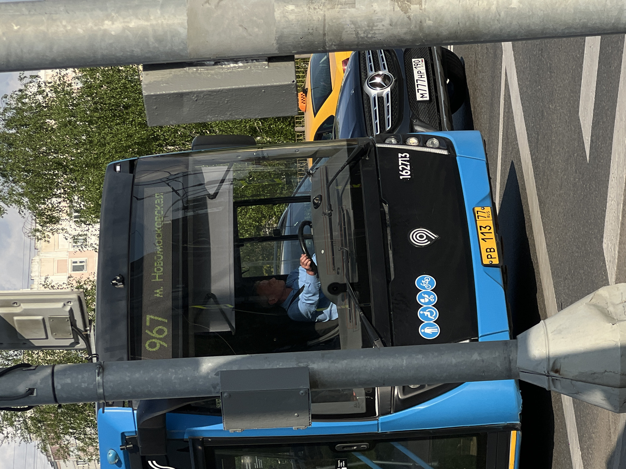 The image captures a blue city bus, identified by its route number 967 displaying "М. Новомосковская" (M. Novomoskovskaya), situated in Moscow, Russia. The bus driver is visible behind the wheel. The bus's license plate reads "PB 113 77 RUS" and the fleet number "162713" is on the side. The image also includes a glimpse of a yellow taxi and a dark-colored Mercedes Benz car with a license plate "M777HP190 RUS" in the background. A grey metal pole obstructs part of the view. The environment appears to be a typical urban street with road markings and greenery.