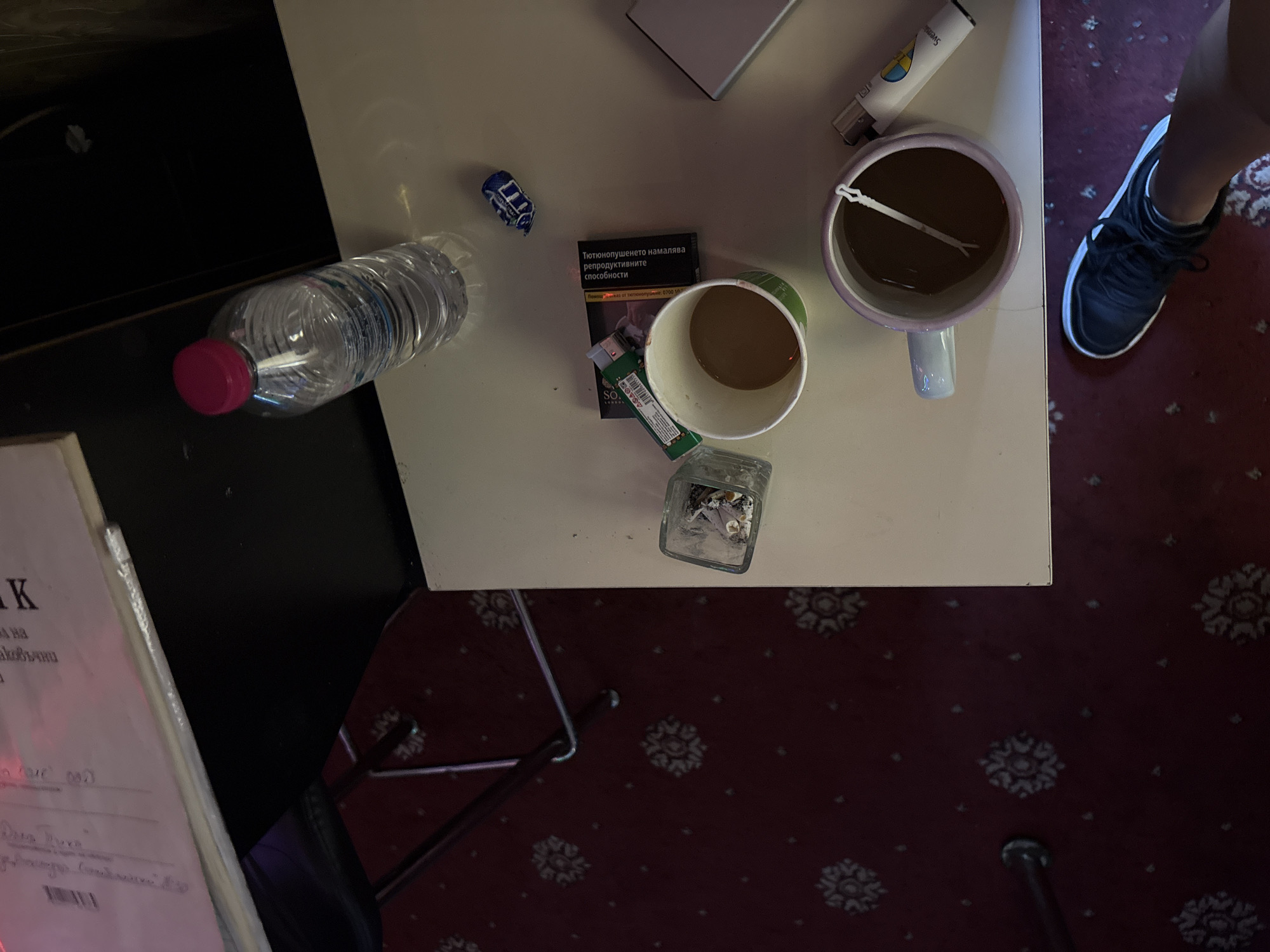 The image depicts a close-up, high-angle view of a table with various items, suggesting a casual setting. In the foreground, a person's leg and foot wearing a black athletic shoe are visible. On the table, a clear plastic water bottle with a pink cap stands to the upper right. Below the bottle, a crumpled blue candy wrapper is present. To the left of the wrapper, a white paper cup contains a dark liquid, possibly coffee, and a green cigarette lighter rests on top of a pack of cigarettes. Further left, a small glass ashtray holds cigarette butts. In the lower right of the frame, a mug with a light purple rim holds a dark liquid and a white stirring stick. A white and yellow lighter is placed next to the mug. The table surface is a light color, and the background shows a dark surface and a patterned red carpet. The scene appears to be indoors, possibly in a residential setting or a casual establishment. Based on the provided information, this scene is located in the city of Karlovo, Bulgaria.