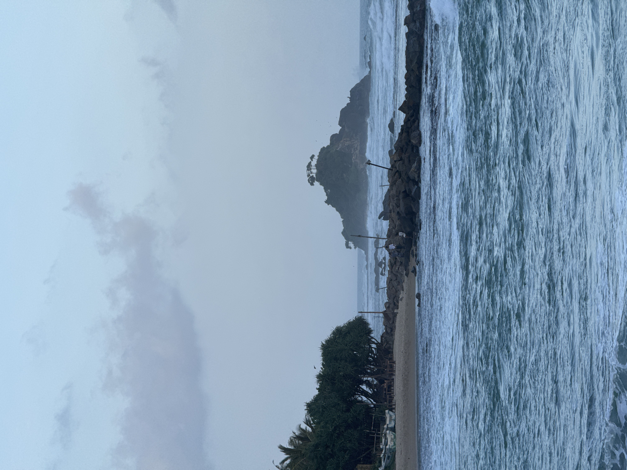 This is a photograph of a coastal scene in Goviyapana, Sri Lanka, likely taken at dawn or dusk due to the soft lighting and muted colors. The foreground is dominated by choppy, turquoise ocean water with white foam cresting on some waves.
In the middle ground, a man-made breakwater constructed from large, dark rocks extends from the right side of the frame towards the left, partially obscuring the view of the shoreline. Several people are visible standing on this breakwater, some looking out at the sea. Beyond the breakwater, a sandy beach is visible, lined with dense, green vegetation including palm trees. Several small fishing boats are pulled up onto the sand near the vegetation. Further out in the ocean, a small, rocky island topped with a cluster of trees stands against the horizon. The sky above is a pale blue, with wispy, light gray clouds scattered across it. The overall impression is one of a serene, yet somewhat wild, coastal landscape.