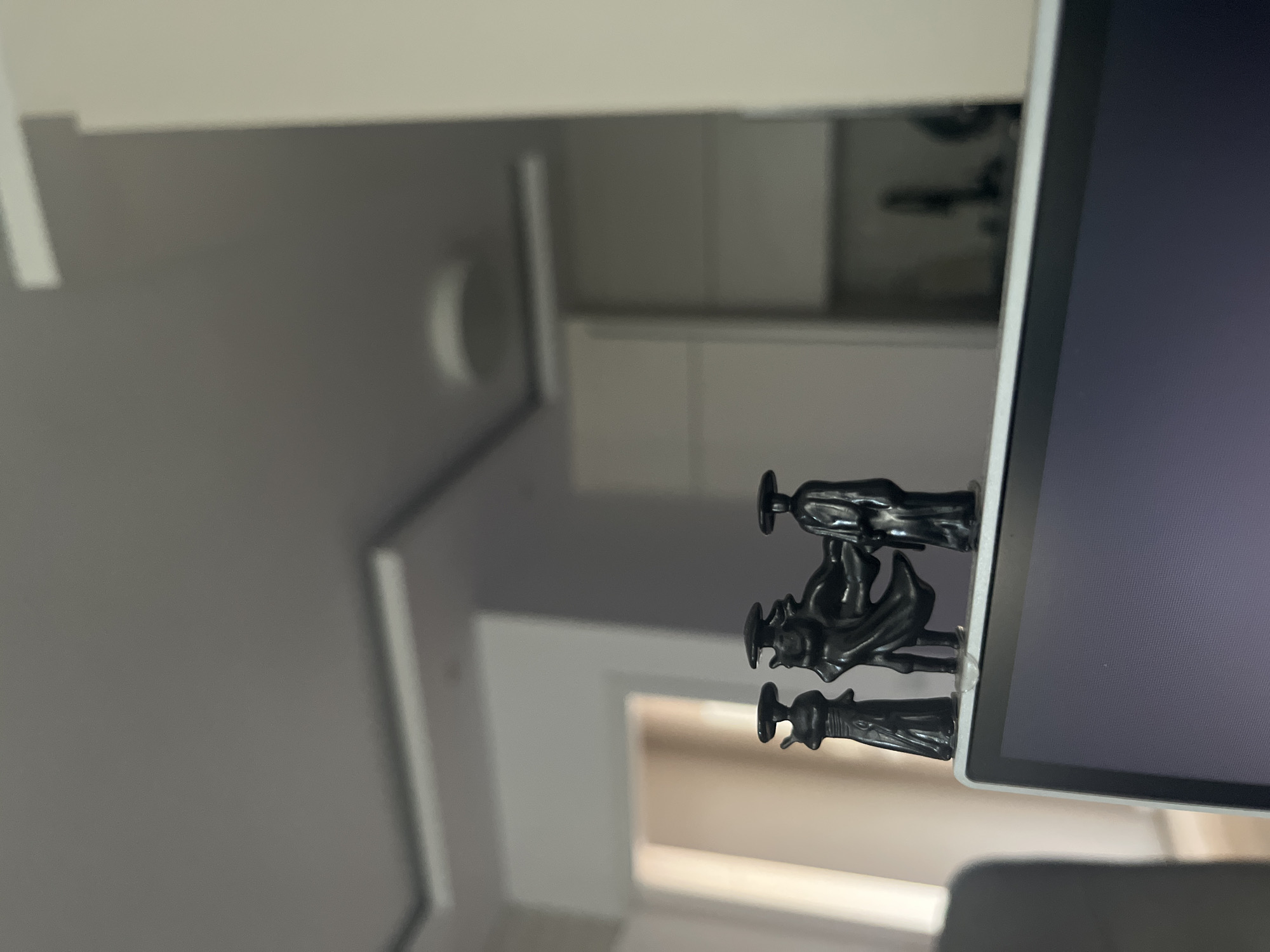 Three black miniature figurines of figures in cloaks and hats stand on the top edge of an open laptop. The figurines are positioned side-by-side, with the middle figurine appearing to be larger or more prominent, possibly in a dynamic pose. The laptop screen is visible in the foreground, displaying a dark, reflective surface. The background is blurred, showing interior architectural details such as a ceiling with recessed lighting, a wall, and what appears to be kitchen cabinetry. The lighting suggests an indoor environment, possibly a home or office. There is no indication of the city of Varna, Bulgaria, in the image.