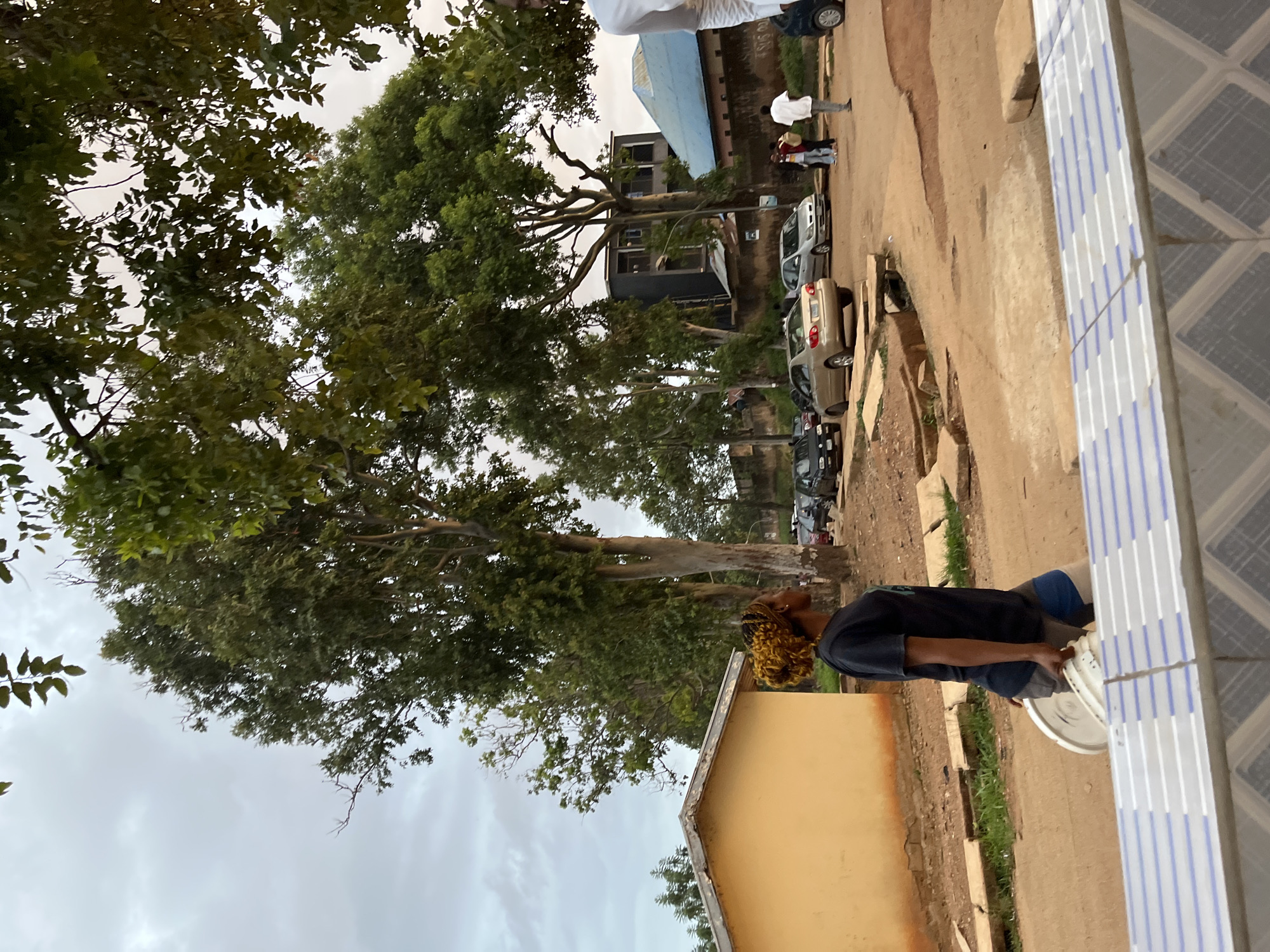 In Jos, Nigeria, on a cloudy day, a woman with bright yellow, curly hair walks across a dirt and gravel road carrying a white bucket. She is wearing a dark t-shirt and shorts. Behind her, a light yellow building stands to the left, and several cars are parked or driving on the road. In the background, there are trees and some buildings, including a multi-story structure and one with a light blue roof. A man in a white shirt walks away from the camera on the right side of the image. The scene appears to be an outdoor area with a mix of residential and possibly commercial structures.