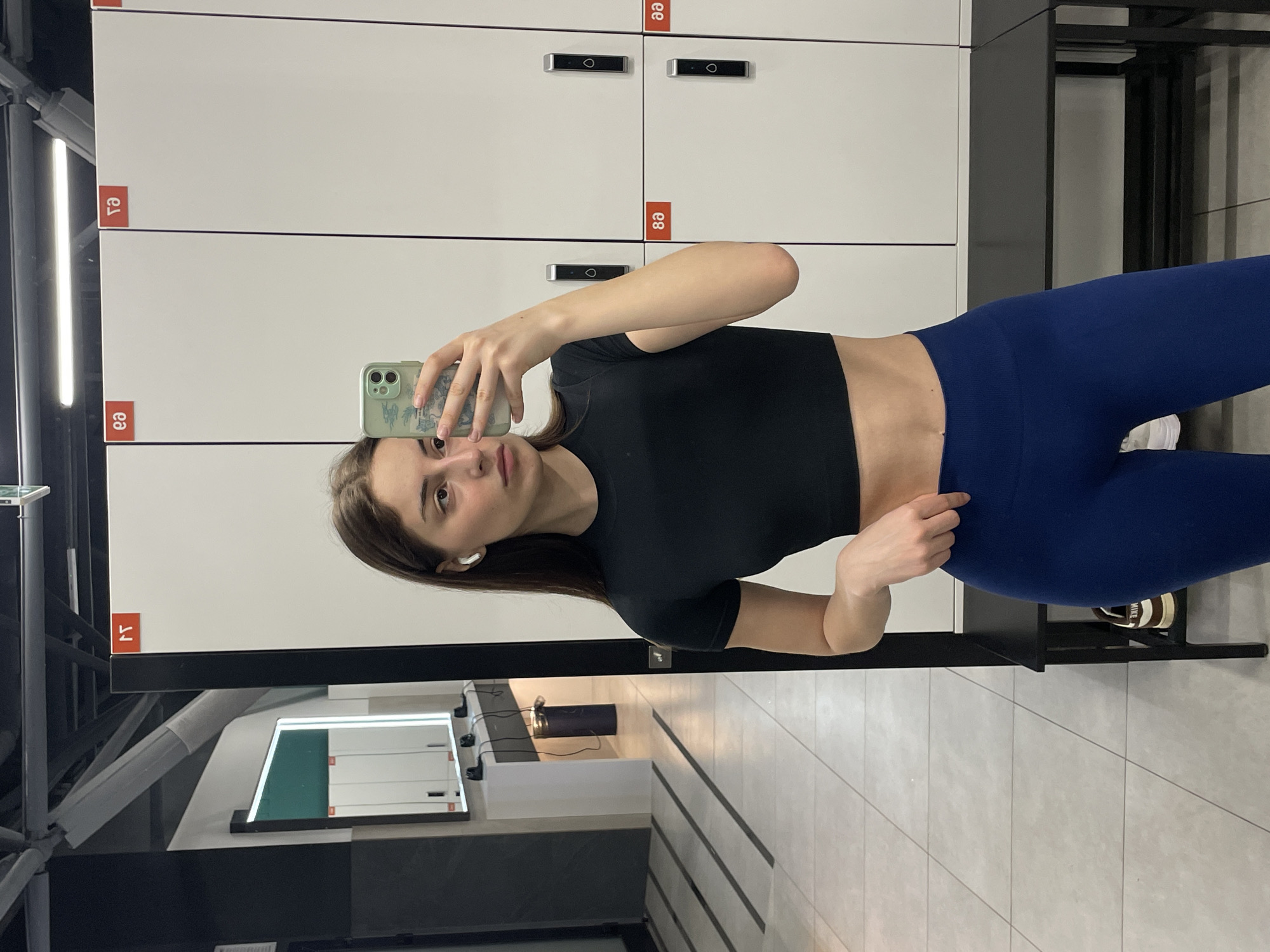 A woman stands in what appears to be the changing room of a gym in Moscow, Russia, based on the Cyrillic lettering on the locker numbers. She is taking a selfie with her smartphone. She is wearing a black crop top and blue leggings. Her hair is brown, and she has an earbud in her left ear. The background shows rows of white lockers with electronic keypads. The floor is tiled. Part of a mirror or reflective surface is visible in the lower left, showing what seems to be a lighted display or sign.