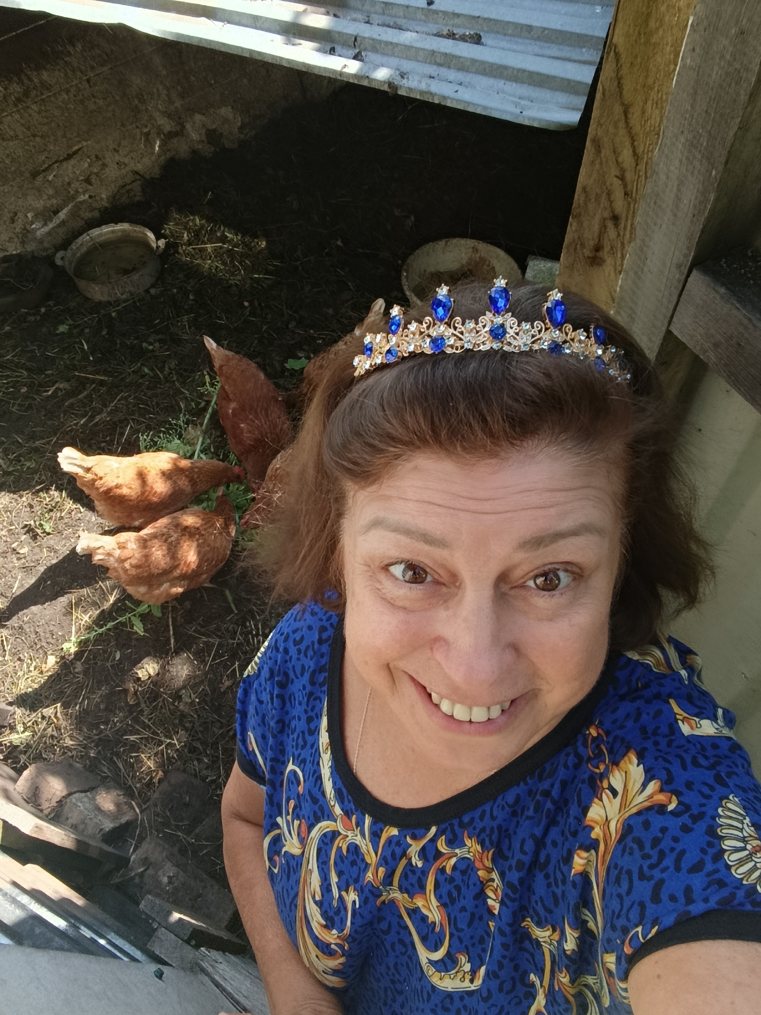 A woman wearing a gold crown adorned with blue jewels and a blue patterned shirt smiles for a selfie. She is outdoors, in what appears to be a farmyard or rural setting. In the background, three brown chickens are pecking at the ground. To the left, there is a dark, possibly muddy area with a shallow, round trough. To the right, there is wooden fencing and what looks like the corrugated metal roof of a structure. The ground is covered with dry grass and dirt. The scene suggests a casual and somewhat whimsical moment, possibly on a farm located in or near the city of Верея, Russia, given the context provided.