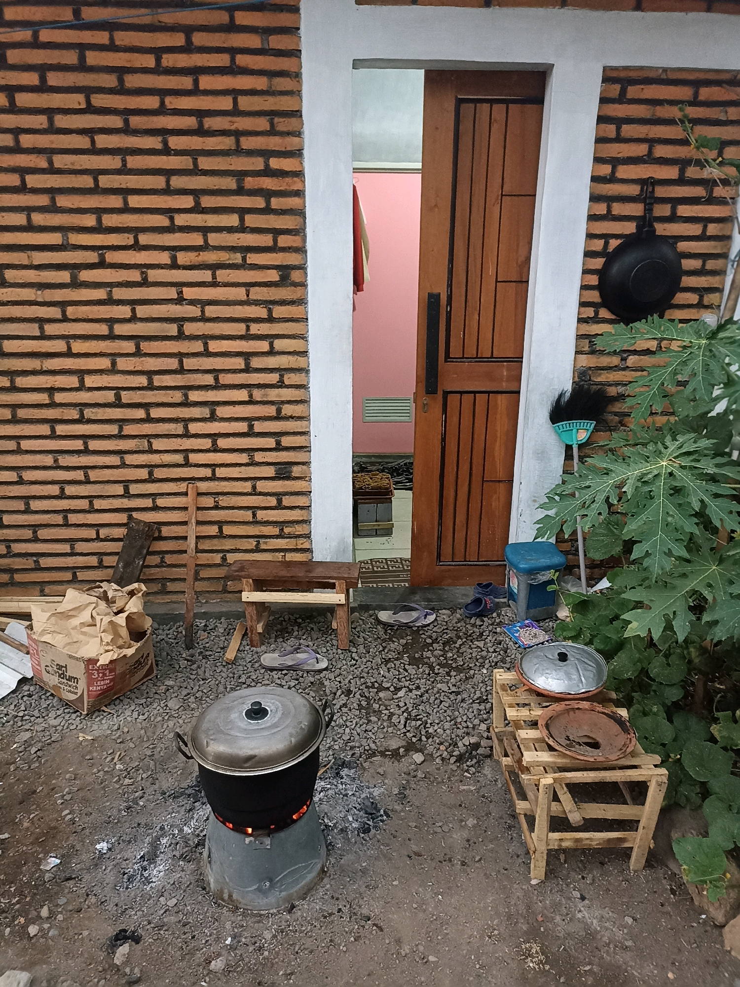 The image depicts an outdoor cooking scene in Mojopanggung, Indonesia. A black pot with a silver lid sits atop a makeshift stove, with glowing embers visible beneath it. The stove appears to be a repurposed metal container. In front of the stove, to the right, is a small, rustic wooden stand holding two clay pot lids. To the left of the stove, a crumpled cardboard box with some text is placed on the gravelly ground. A wooden stool with a plank seat is positioned behind the box. Near the stool, a pair of flip-flops rests on the ground. Further back, a wooden door with vertical planks is ajar, revealing a pink-walled interior. A black frying pan hangs on the brick wall to the right of the door. A broom with a green handle and black bristles leans against the wall next to the door. A blue trash can is situated near the broom. A large, leafy green plant, possibly a papaya tree, is growing to the right of the scene. The ground is covered in gravel and dirt, and the wall is constructed of orange bricks with dark mortar. The overall impression is one of a utilitarian outdoor cooking area.