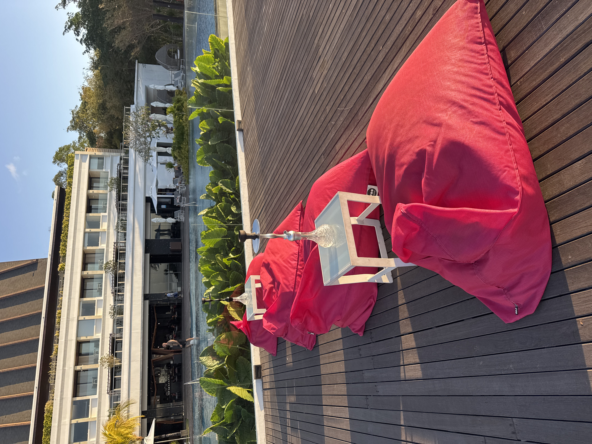 This is a medium shot, taken at eye level, of a row of pink beanbag chairs on a wooden deck overlooking a swimming pool and a resort building, likely at sunset in Kuta Selatan, Indonesia. The resort building, seen in the background, is a multi-story white structure with dark-framed windows and balconies. Lush green foliage separates the deck from the pool, which is blue and reflects the sky. Two hookahs with ornate glass bases and metal stems are placed on small white tables in front of the beanbag chairs. The deck is made of dark wood planks running horizontally. The lighting suggests late afternoon or early evening, with long shadows cast across the deck. No people are actively engaged in any interactions, but one person is visible in the pool in the mid-ground.