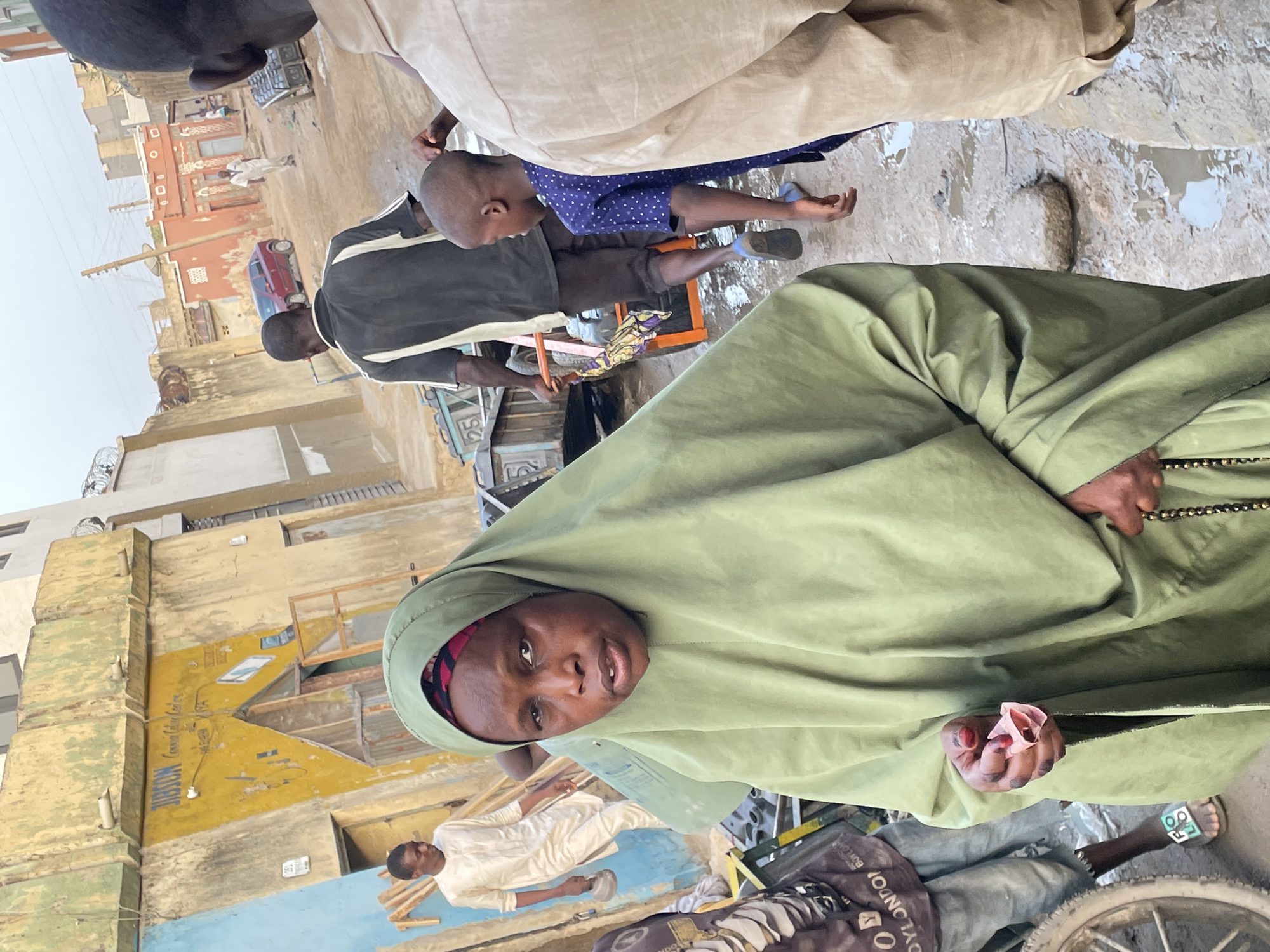 A daytime outdoor scene shows a woman in a green headscarf and dress holding a handful of money in her left hand and prayer beads in her right. The background contains multiple people, including a man in a black-and-white shirt, a boy in a blue shirt, and a man in a beige outfit. The location is an urban street with buildings and a painted advertisement on a wall. The city is Unknown, Unknown.