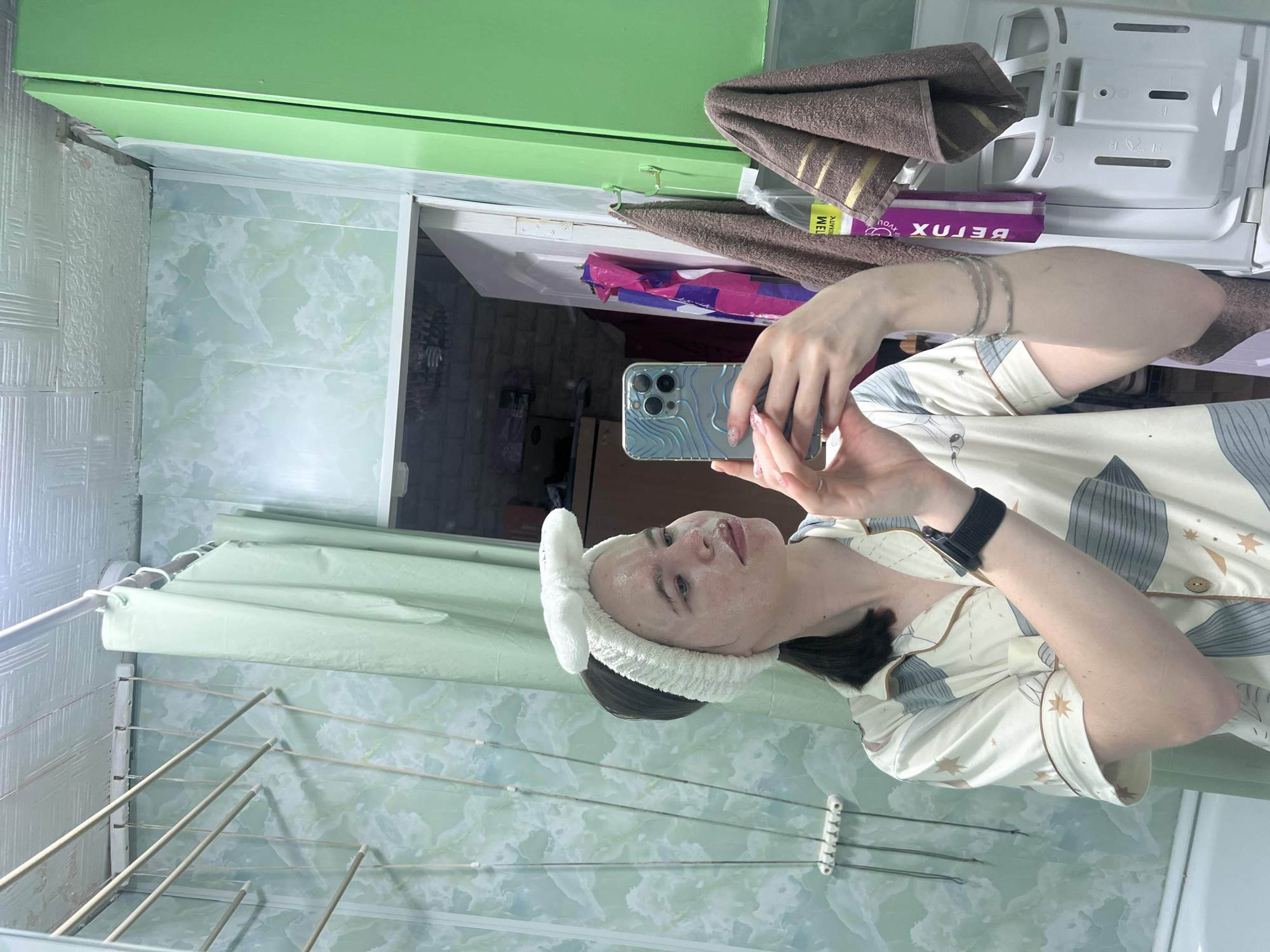 A woman is taking a selfie in a bathroom. She is wearing pajamas and a plush headband. The bathroom has green and white marbled wallpaper, a clothes drying rack, and a cabinet above the toilet. The scene is in an unspecified location in the city of Chaikovsky, Russia.