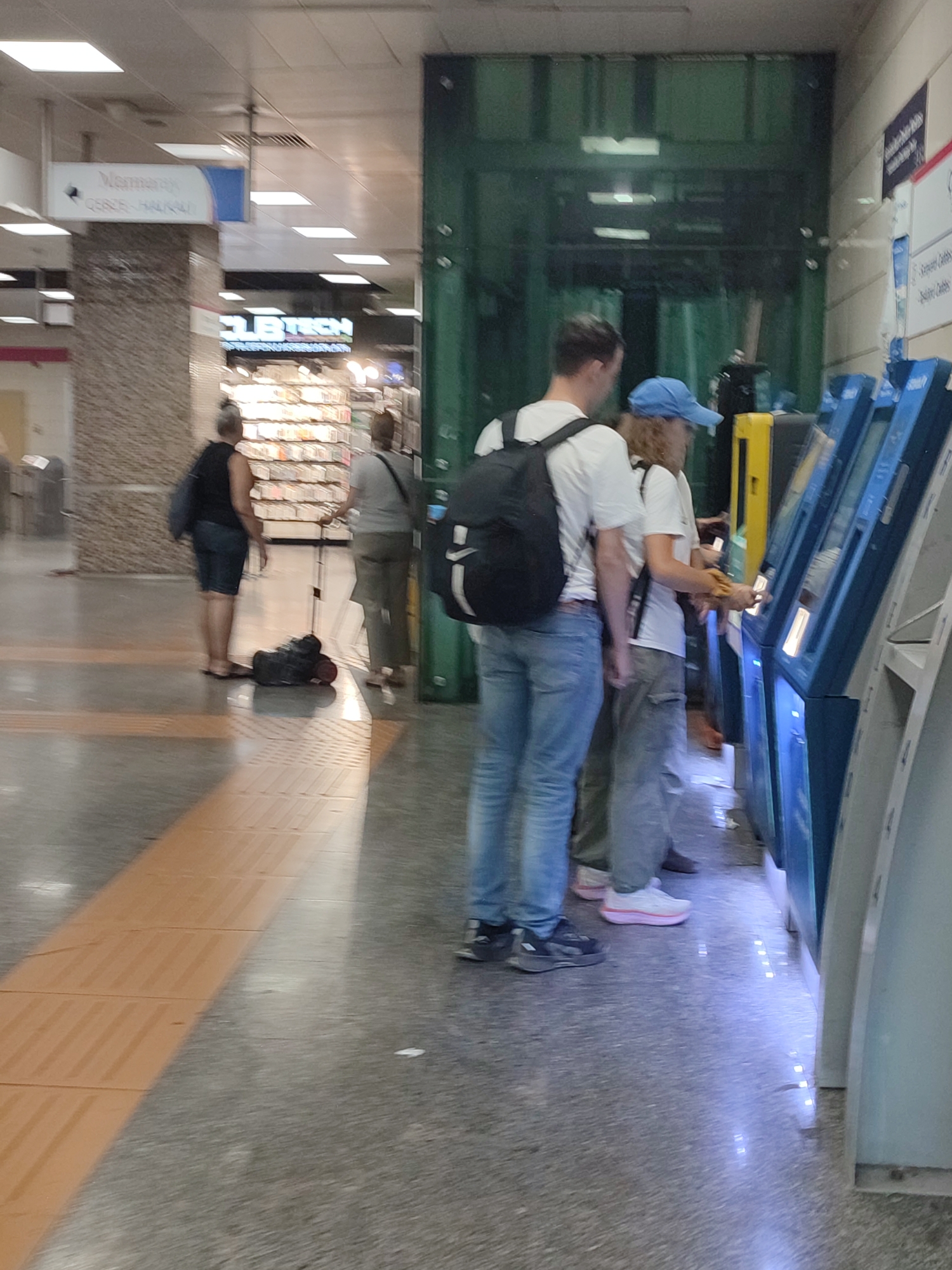 In Kadıköy, Turkey, at a transit station, a group of individuals are observed interacting with ticket machines. A man wearing a white t-shirt and blue jeans, carrying a black backpack, stands to the left of the frame, facing a row of blue ticket kiosks. To his right, a woman with curly hair, wearing a blue cap and a white t-shirt, is also by the ticket machines, with her hand near the screen. Another woman, further back and to the left, in a black sleeveless top and denim shorts, is seen with a dark bag slung over her shoulder, with a black suitcase on the floor beside her. A person in a grey t-shirt and pants is visible in the background, also near the ticket machines. The environment appears to be an indoor public space with tiled flooring and overhead lighting. The ticket machines are illuminated, suggesting they are in operation.
