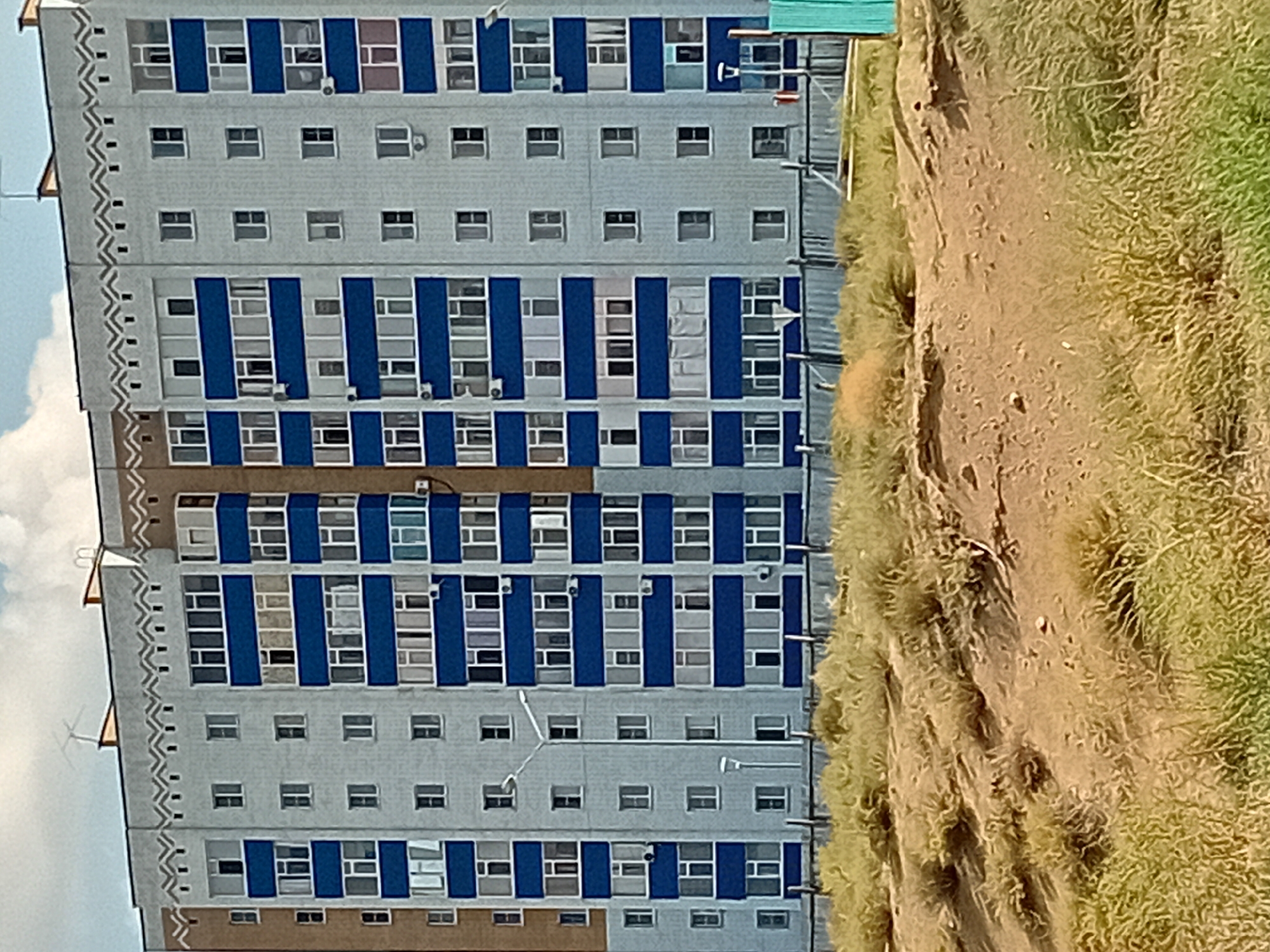 The image shows a multi-story apartment building in Semey, Kazakhstan. The building has a light gray facade with horizontal bands of dark blue. Each floor has multiple windows and some balconies. The building is several stories high, with an antenna visible on the roof. In front of the building is a fence and a grassy, uneven terrain with some dry vegetation and sparse bushes. The sky above is partly cloudy. The architecture and the surrounding environment suggest a residential area in a city, likely a block of flats.