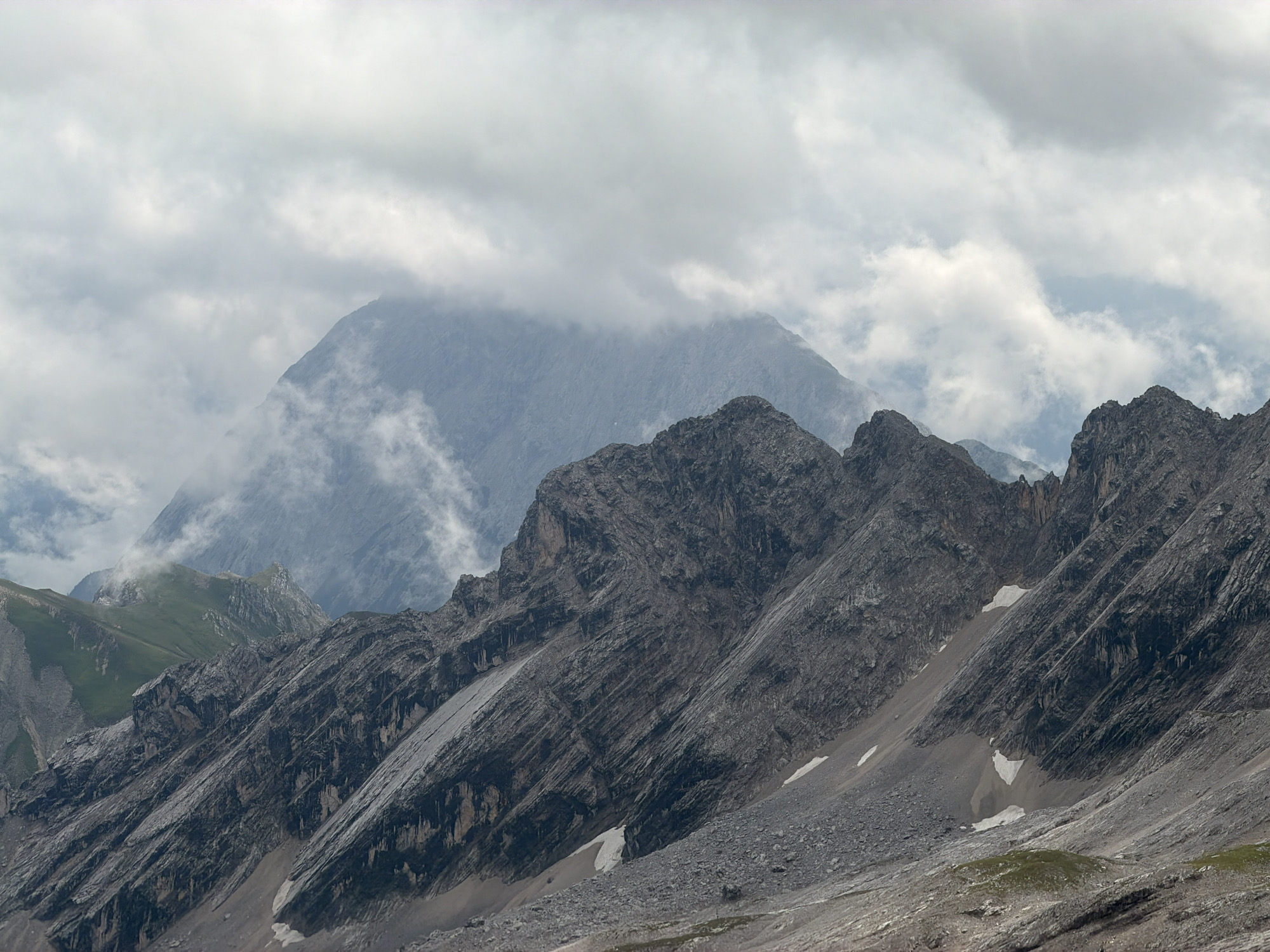The image depicts a mountainous landscape under a cloudy sky. The foreground features a steep, rocky slope with some patches of snow and scattered grey scree. The midground is dominated by rugged mountain ridges with sharp, angular peaks, also covered in rock and scree. In the background, a larger, more distant mountain peak is partially obscured by mist and clouds, creating a sense of depth and atmosphere. To the left of the background peak, a verdant green mountain slope is visible, contrasting with the grey rock. The sky is filled with an overcast of grey and white clouds, with hints of blue sky peeking through in some areas. There are no people or man-made structures visible in the image. The scene is likely located in the Bavarian Alps, given the characteristic alpine topography. Without further information, pinpointing the exact location within Garmisch-Partenkirchen is not possible, but the image captures the essence of the high alpine environment surrounding the town.