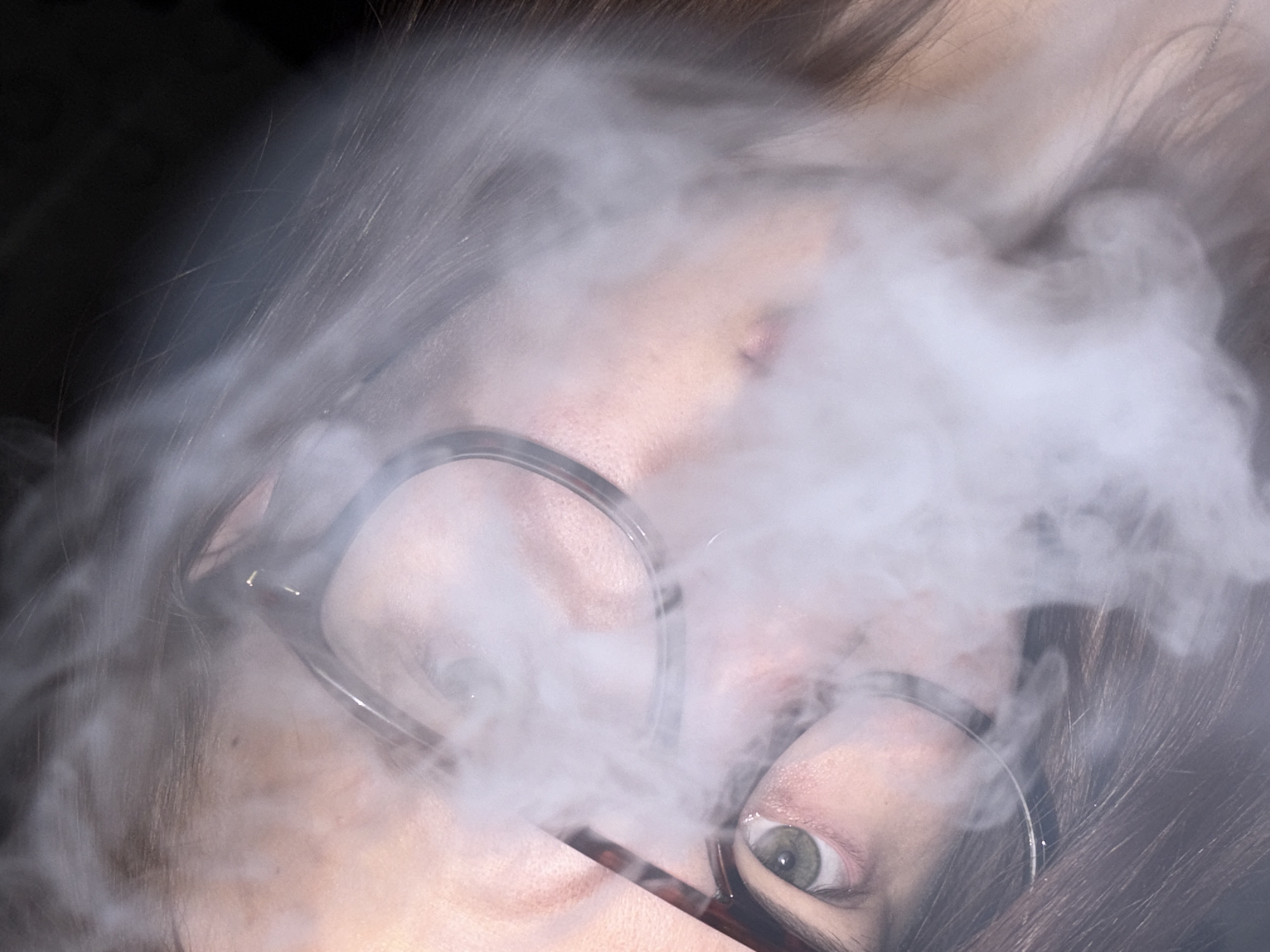The close-up, rotated image captures a person's face partially obscured by dense, white smoke or vapor. The subject has dark hair, visible around the edges of the frame, and wears eyeglasses with dark, possibly tortoiseshell, frames. One eye, specifically the person's left eye (viewer's right), is clearly visible, revealing a light green or hazel iris and a reflective pupil.
The smoke or vapor swirls prominently across the center of the frame, covering the area around the nose and mouth, and softening the facial features. The skin tone appears light. The background is dark and indistinct, suggesting an indoor setting or low-light conditions. The primary activity depicted is the interaction of the person with the smoke, likely exhaling vapor from vaping or smoke. The visible eye conveys a neutral or contemplative expression. No specific location cues, time of day indicators beyond low light, or visible text are present in the image.
