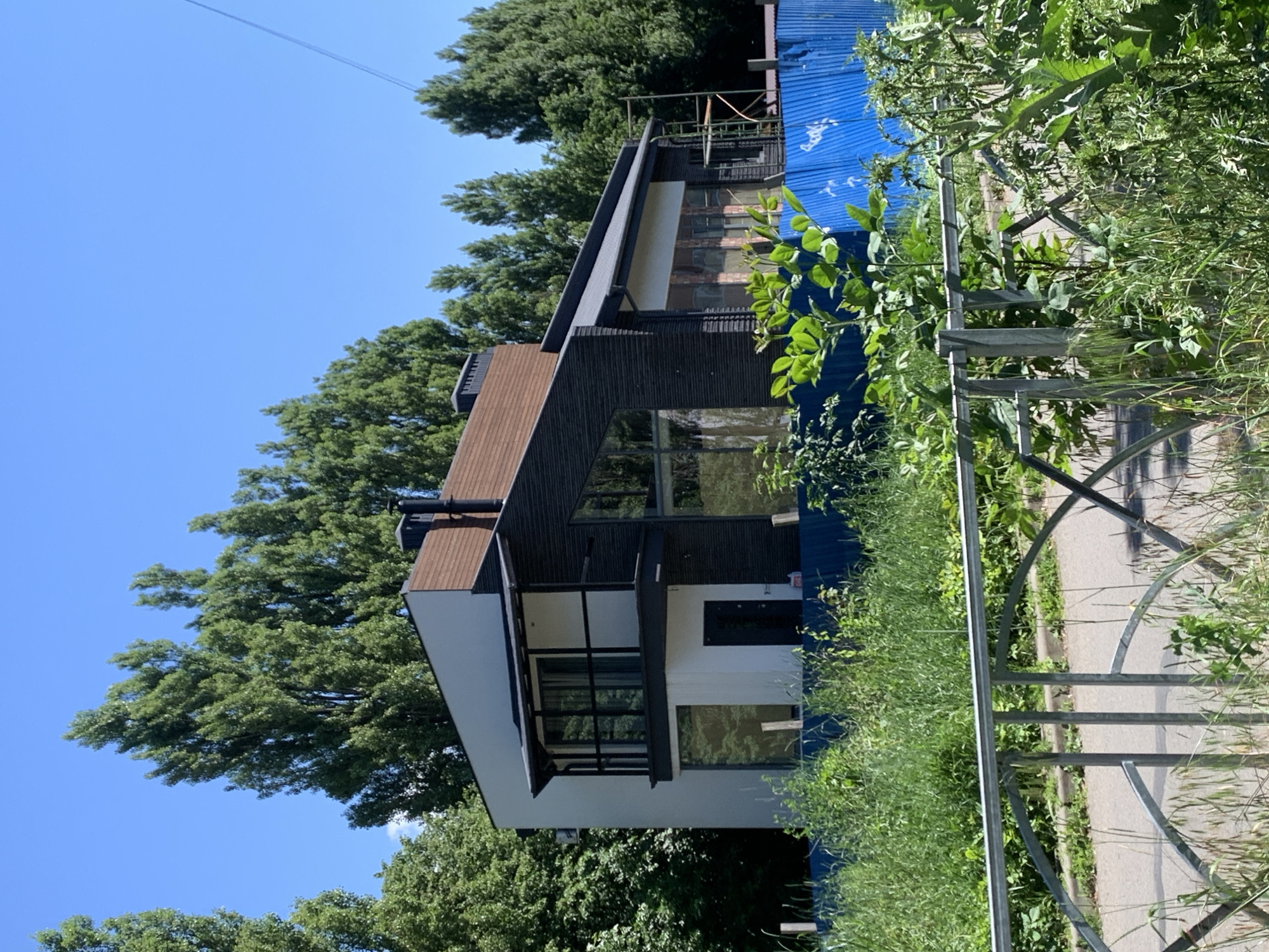 The image, taken from a low angle in Stavropol, Russia, features a modern house with dark siding and large windows. The house has a prominent angled roof covered in brown shingles. Adjacent to the house, a bright blue corrugated metal structure is visible, possibly a temporary covering or construction element. Lush green trees surround the property, with some foliage encroaching on the frame from the right side, obscuring a portion of a metal fence. The sky is clear and bright blue, suggesting a sunny day. The overall scene appears to be set in a residential area with significant natural landscaping. No people are visible, and the image focuses on the architecture and its surroundings. There is no discernible text within the image.