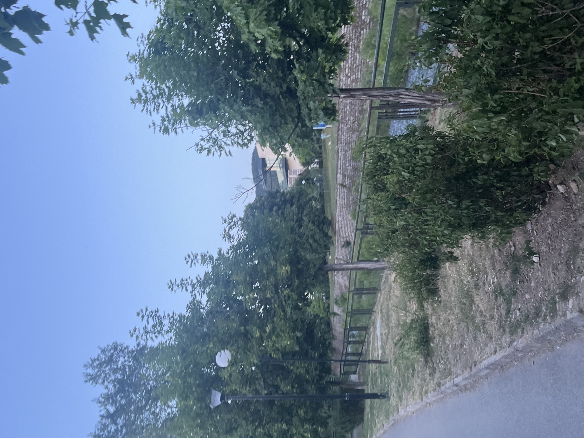 The image shows a park or green area in Blagoevgrad, Bulgaria. A paved walkway runs along the right side of the frame. A metal fence borders the walkway, separating it from a grassy area that leads to a brick-paved section and a water feature. Trees and bushes line the walkway and obscure some of the view. A building is visible in the distance. A street lamp is located along the path. The sky is visible in the upper left corner.