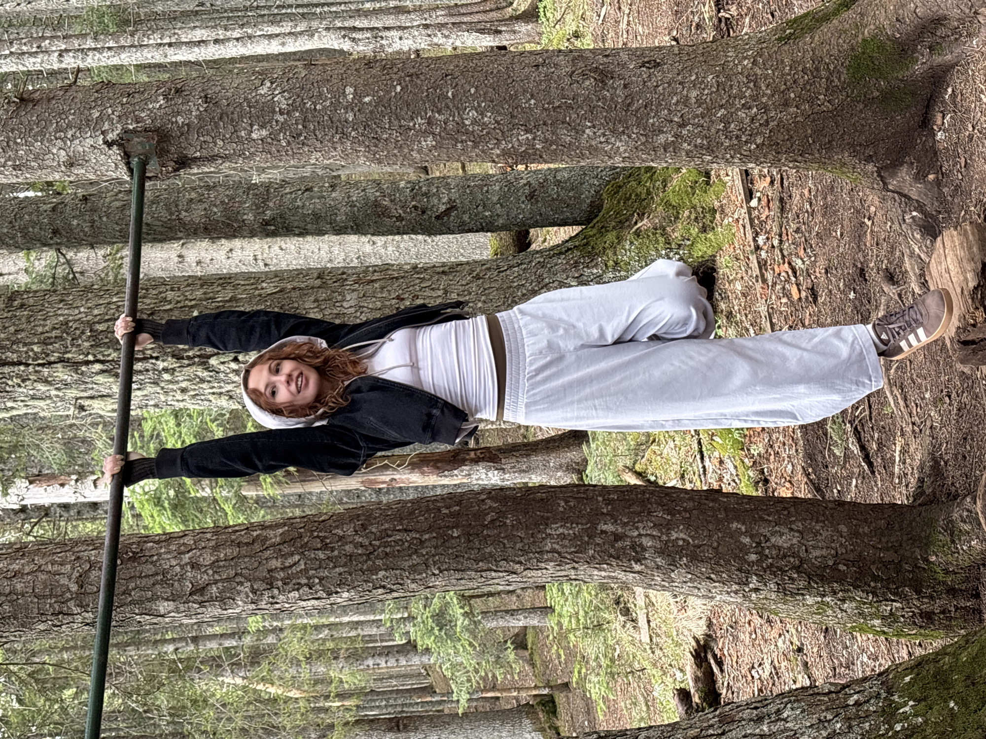 A young woman stands in a forest, holding onto a vertical pole with both hands. She wears light grey sweatpants, a white tank top, and a black jacket with a grey hoodie pulled up. Her reddish-brown hair is visible around the hood. She is smiling and looking towards the camera. The background is a dense forest with many tall trees. The ground is covered in fallen leaves and moss.