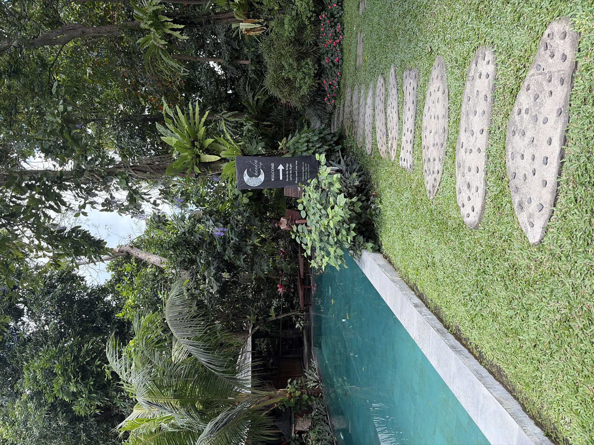 The image depicts a lush, tropical garden setting in Mengwi, Indonesia, featuring a swimming pool and a stepping stone pathway. The pool, with its clear turquoise water, is bordered by a concrete edge and lush green grass. To the left of the pool, palm trees and dense foliage create a secluded atmosphere, with a glimpse of a wooden structure in the background. A stone pathway, made of large, irregular stepping stones embedded with dark pebbles, winds through the well-maintained lawn, leading towards the right of the frame. On the right side, a tall black sign with a crescent moon logo and the text "Chandra Yoga" and "WELCOME" stands prominently, indicating the location. Beside the sign, a woman is seated on a wooden bench, looking at her phone. Various tropical plants, including ferns and flowering bushes with pink blossoms, fill the background, contributing to the serene and natural ambiance. The overall scene conveys a sense of tranquility and resort-like appeal.