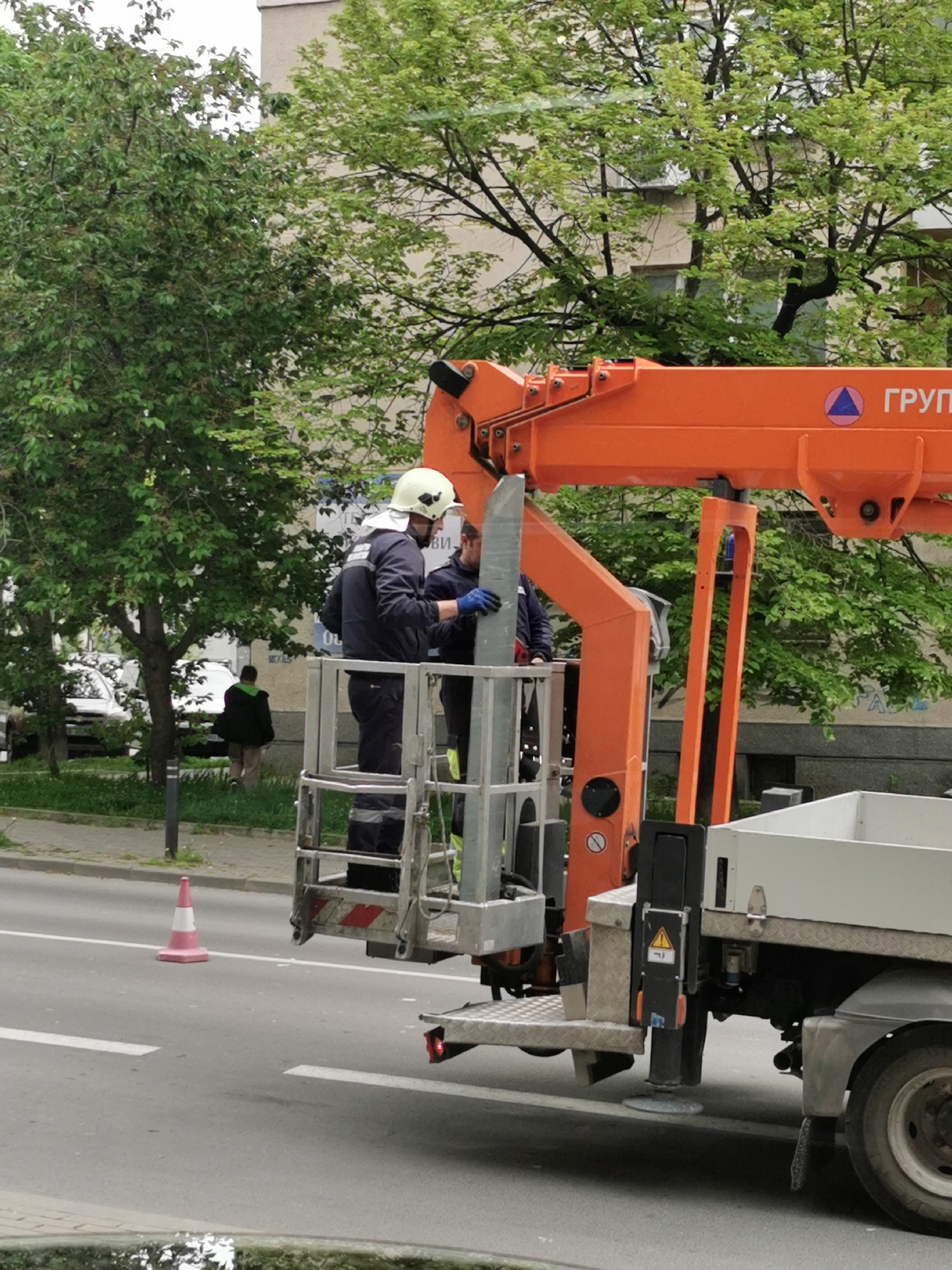 The image depicts two workers in a cherry picker on a street in Varna, Bulgaria. They are wearing dark clothing and hard hats, and appear to be working on something together. The cherry picker is mounted on a truck parked on the side of the road. A traffic cone is placed on the road near the cherry picker. The background shows a city street with trees, buildings, and cars.