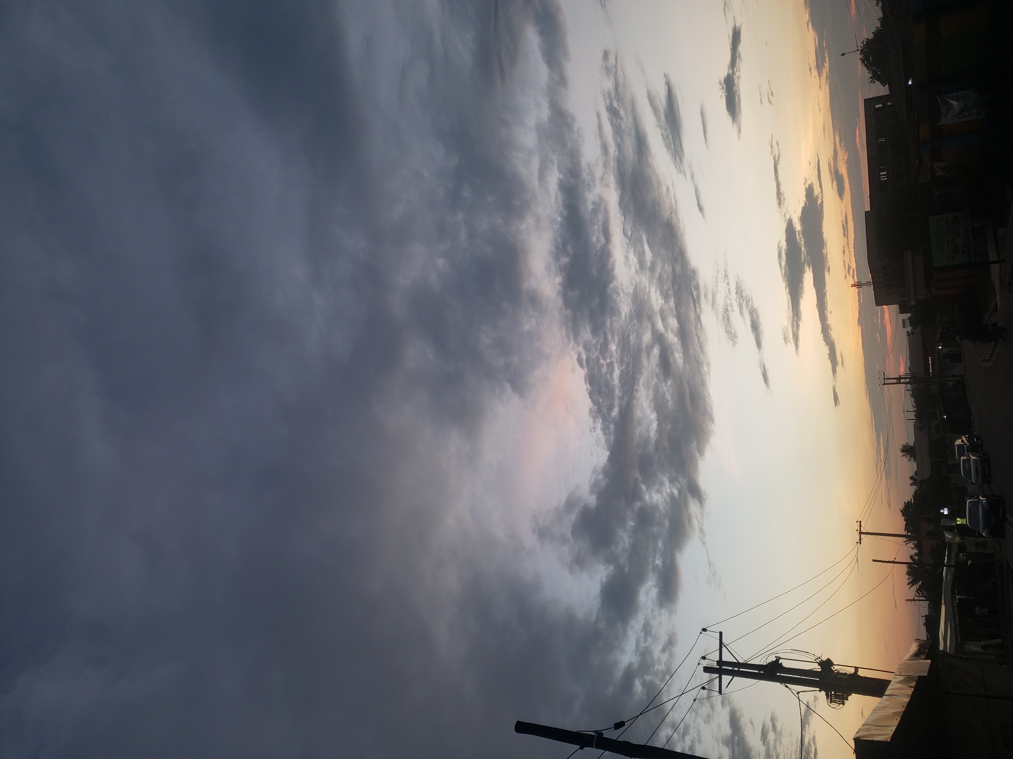 The image depicts a street scene in Narayi, Nigeria, during dusk. The sky is filled with dramatic clouds, ranging in color from dark gray to lighter shades with hints of pink and orange near the horizon, indicating the setting sun. In the foreground, a utility pole with numerous wires stands prominently on the left side of the image. The road, partially visible, has several vehicles, including cars and what appears to be a motorcycle, driving along it. Along the sides of the road, there are buildings and structures, some of which appear to be shops or market stalls, with their silhouettes darkened against the brighter sky. The overall impression is of a bustling urban environment at the end of the day, with the natural beauty of the twilight sky contrasting with the man-made structures and infrastructure.