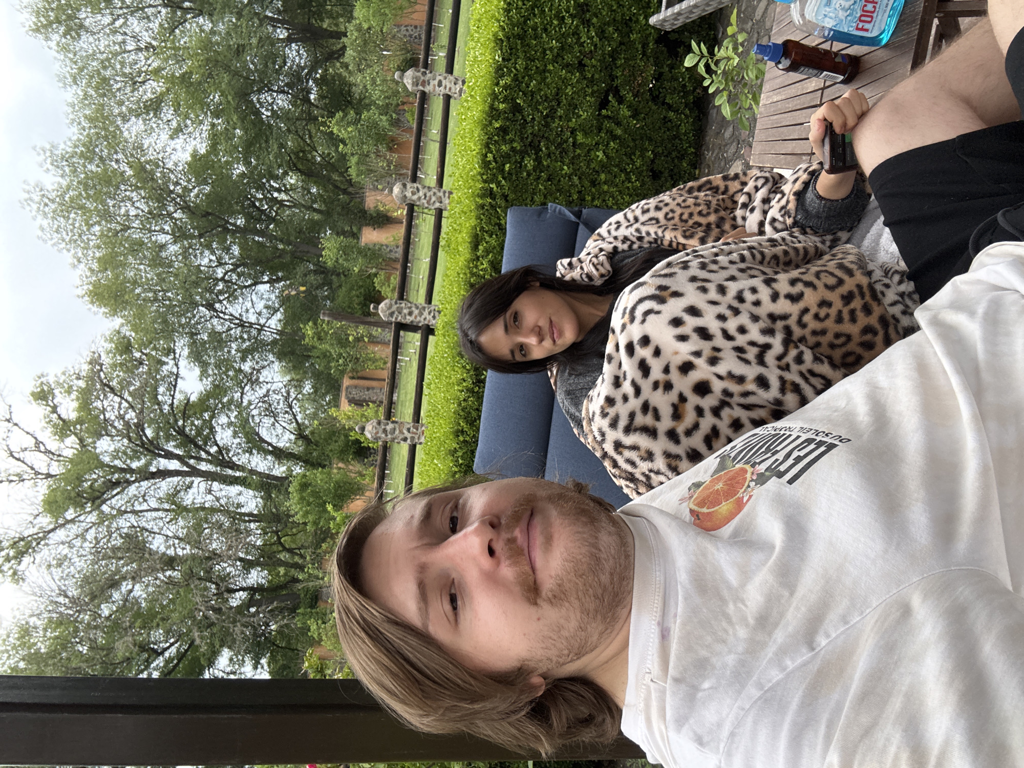 Two people are outdoors in Cañada de Madero, Mexico. A man is holding the camera in a selfie pose wearing a white t-shirt. A woman is sitting on a blue cushion, covered by an animal print blanket. A table is next to her with a blue bottle and a brown bottle on top. Behind them is a green bush and a stone fence. Trees are visible in the background. The sky is overcast.