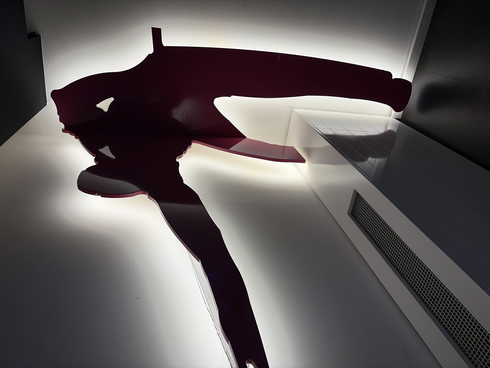 The image depicts a stylized, dark red metal sculpture of a skier mounted on a white wall with integrated backlighting. The sculpture is positioned above a white architectural element with a dark gray grille. The scene appears to be indoors. Based on the visual information, pinpointing the exact location in Budva, Montenegro, is not possible without further context or identifying features.