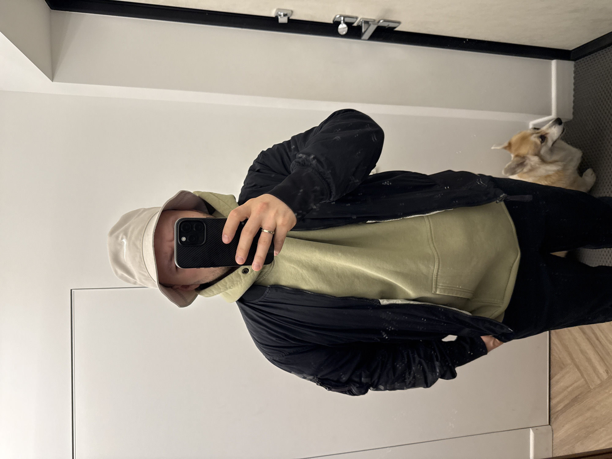 A person takes a selfie in a bathroom. The person is wearing a beige bucket hat, a light green hoodie, and a black jacket. They are holding a smartphone. A tan corgi is visible in the corner of the room. The bathroom has white walls and light wood-patterned flooring. The ceiling is white with a black trim. There are two lights above a mirror. The location is not visible.