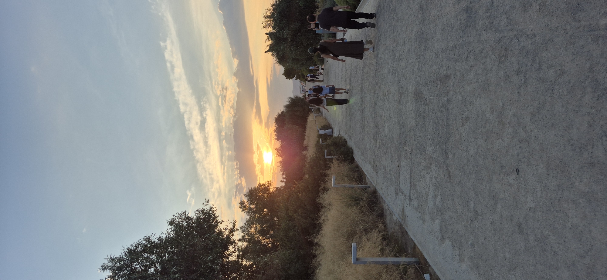 The image depicts a scenic pathway at sunset, located in the Stavros Niarchos Foundation Cultural Center in Kallithea, Greece. The pathway is paved with a light-colored, textured material and is lined on the left with dry, tall grasses and scattered, modern-style lamps. On the right side of the path, there are dense green trees and shrubs. A group of people are seen walking along the pathway, mostly from behind, in the direction of the sunset. The sun is low on the horizon, casting a warm, golden glow and creating a dramatic sky with streaks of orange, pink, and blue clouds. The overall impression is one of a peaceful evening stroll in a well-maintained urban green space.
