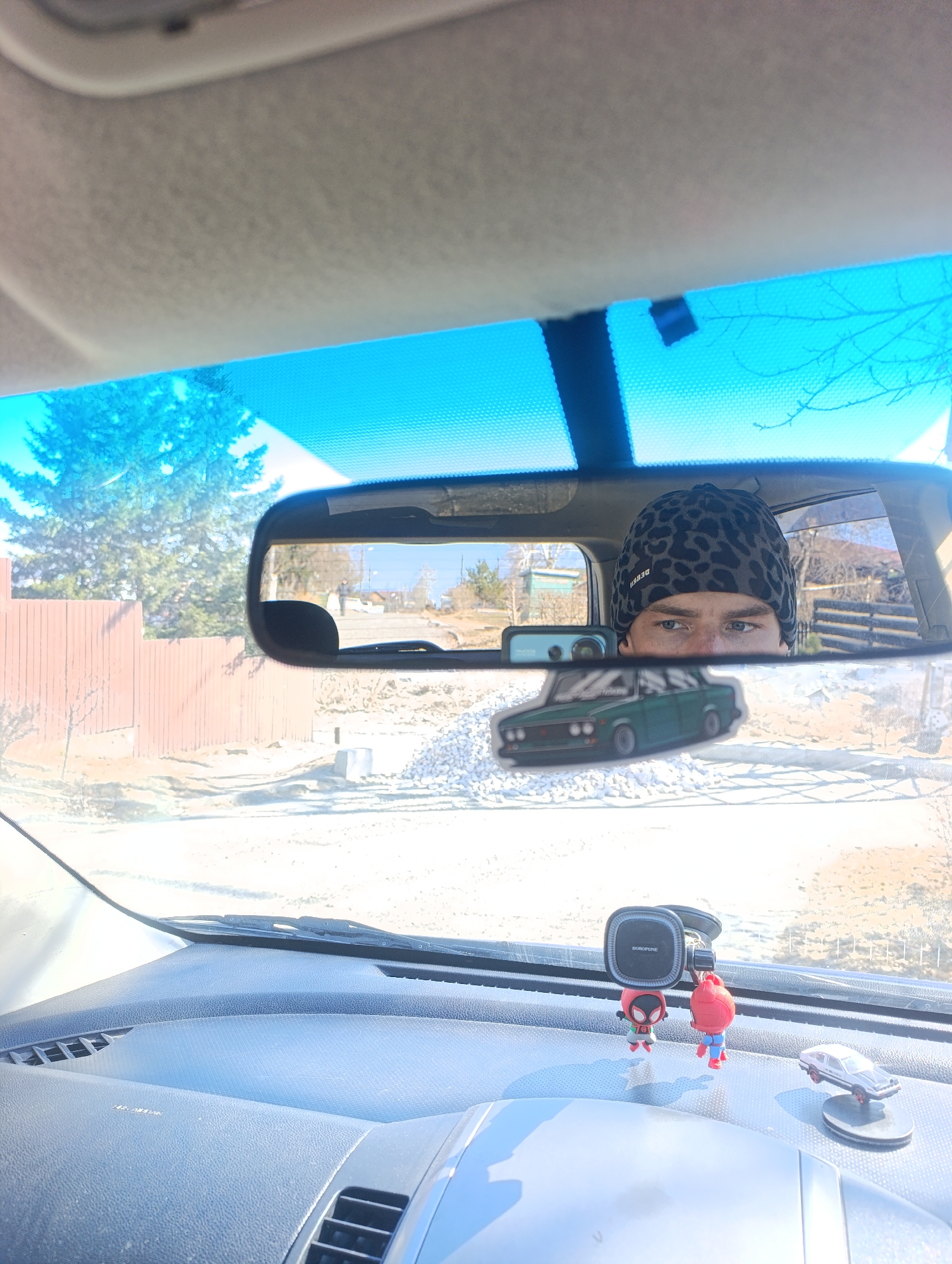 The image, taken from the driver's seat of a car, shows a view through the rearview mirror, reflecting a person wearing a spotted beanie, a street scene, and a sticker of a green car. The reflection includes a view of a street, partially covered with white material appearing to be gravel or construction debris, a brown wooden fence, and some sparse trees in the background. The car's dashboard is visible, featuring a phone mount and two small Spiderman figures and a toy car. Based on the visual information, it is not possible to confirm the exact location in the city of Krasnoyarsk, Russia.