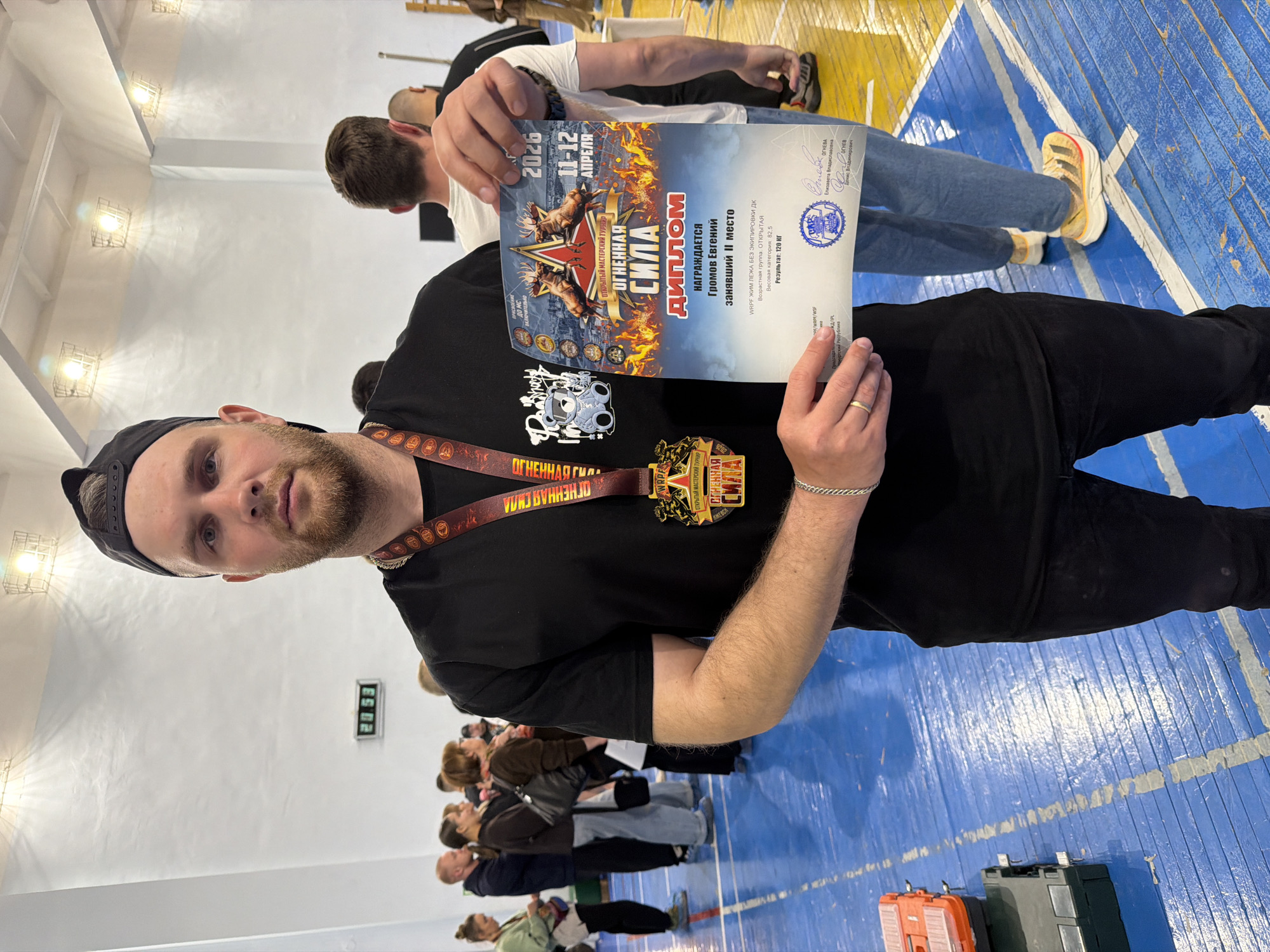 A man is holding a certificate and wearing a medal with the same logo, both displaying the name "OGNENNAYA SILA," likely a competition he participated in. The certificate states the competition took place in Izhevsk, Russia, from April 11-12, 2026. The man, named Gromov Evgeniy, won 2nd place in the WRPF bench press without equipment. He is wearing a black t-shirt with a cartoon-like design, and a black baseball cap. In the background, a group of people are standing. The environment appears to be a gymnasium with blue and yellow striped flooring. There is a digital clock on the wall displaying the time 20:53.