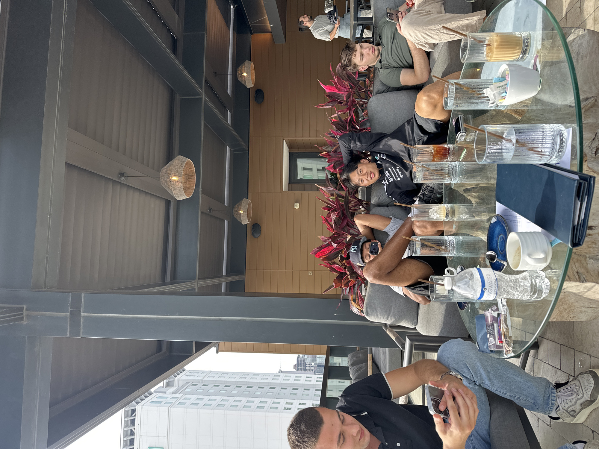 The scene depicts a group of four people sitting in an outdoor seating area, likely a rooftop bar or lounge. The location appears to be Tampa, Florida, suggested by the distant view of buildings resembling the city's skyline. The area is shaded by an overhead structure of metal beams and wicker lampshades.
A round glass table stands in front of the group, covered with several empty drinking glasses, a water bottle, a sugar holder, and a blue folder. The group is sitting on a grey upholstered couch.
One man, clad in a black shirt and blue jeans, is looking at his phone. Two individuals are casually lying back on the couch: one with a black shirt, and the other with a green shirt. A fourth man is sitting farther away.
The setting includes red tropical plants between the couches and the exterior wall.