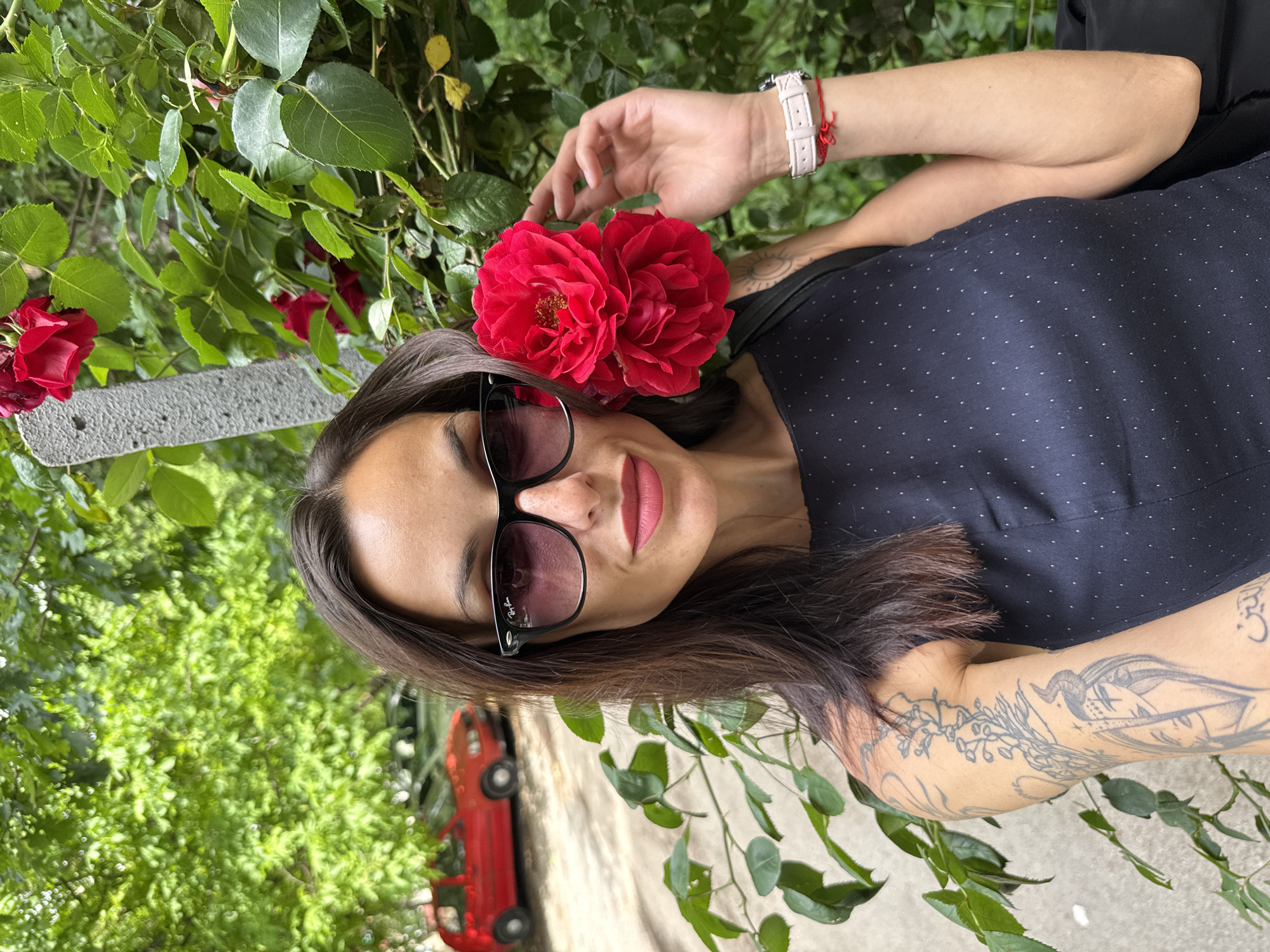 A woman with dark hair, wearing sunglasses and a dark blue top with white dots, is posing outdoors with red roses. She is wearing a white watch with a red string. A red car is visible in the background. The location is Pleven, Bulgaria.