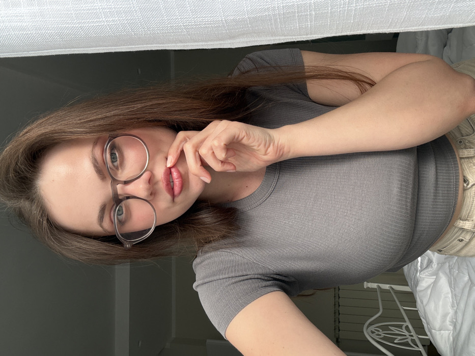 A single female subject is depicted in a close-up, waist-up view within an indoor residential setting in Scafati, Italy. She possesses long, light brown hair and wears clear-framed glasses. Her attire consists of a short-sleeved, ribbed grey top and light-colored pants. Her left hand is raised, with the index finger gently touching her lower lip. The subject is looking directly towards the camera. In the background, to the left, a white metal bed frame with decorative elements and a white radiator are partially visible. To the right, a sheer white curtain partially obscures the background. Lighting appears to be natural, primarily originating from the right side of the frame.
