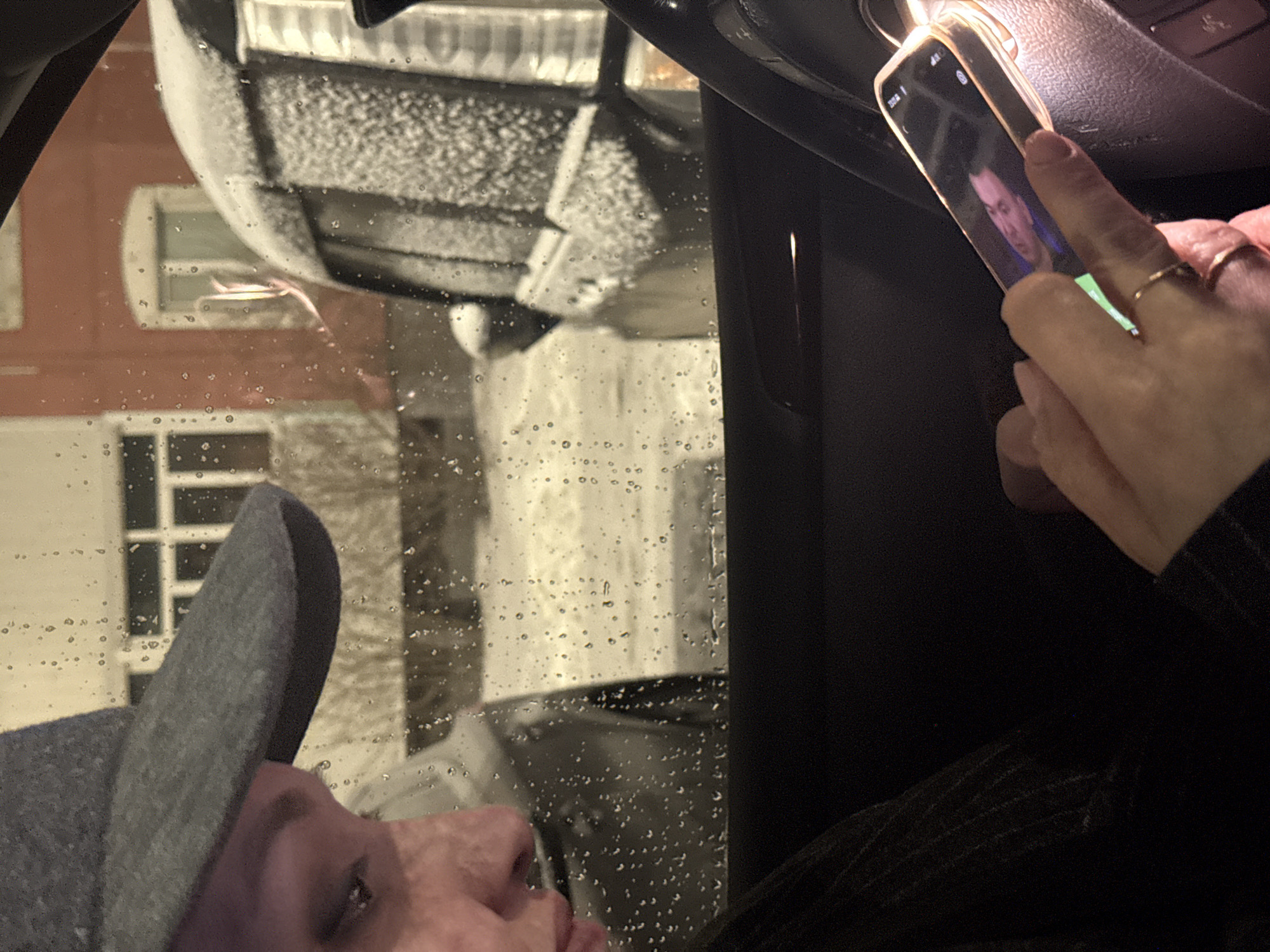 A person inside a car is holding a smartphone displaying an image of a male. The person is wearing a hat and a ring on their finger. The car's windshield is wet, and through it, a building covered in snow can be seen. The location is not discernible from the image. There is no verifiable evidence of Nizhnevartovsk, Russia.