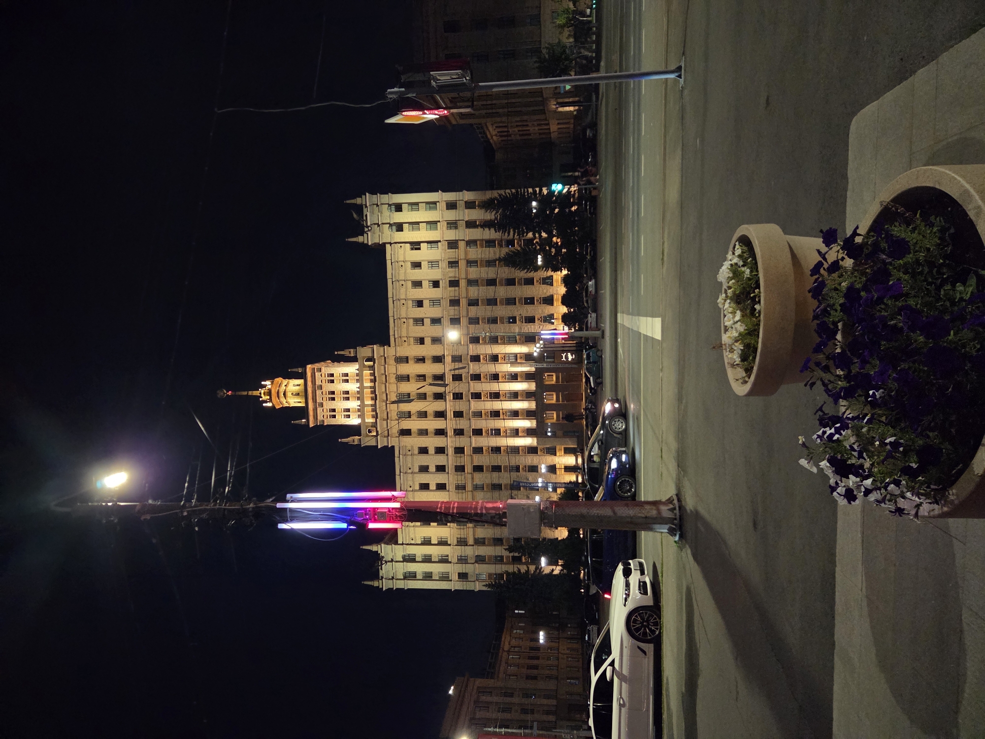 At night, a large, illuminated building with a distinctive spire stands prominently in Chelyabinsk, Russia. The building is a multi-story structure with numerous windows, many of which are lit. In the foreground, a wide street is visible, with several cars driving or parked. A tall lamppost with colorful lights is positioned on the left side of the street. On the right side of the street, a traffic light and another lamppost are present. Two large planters containing flowers are located in the lower right corner of the image. The sky is dark, indicating nighttime.