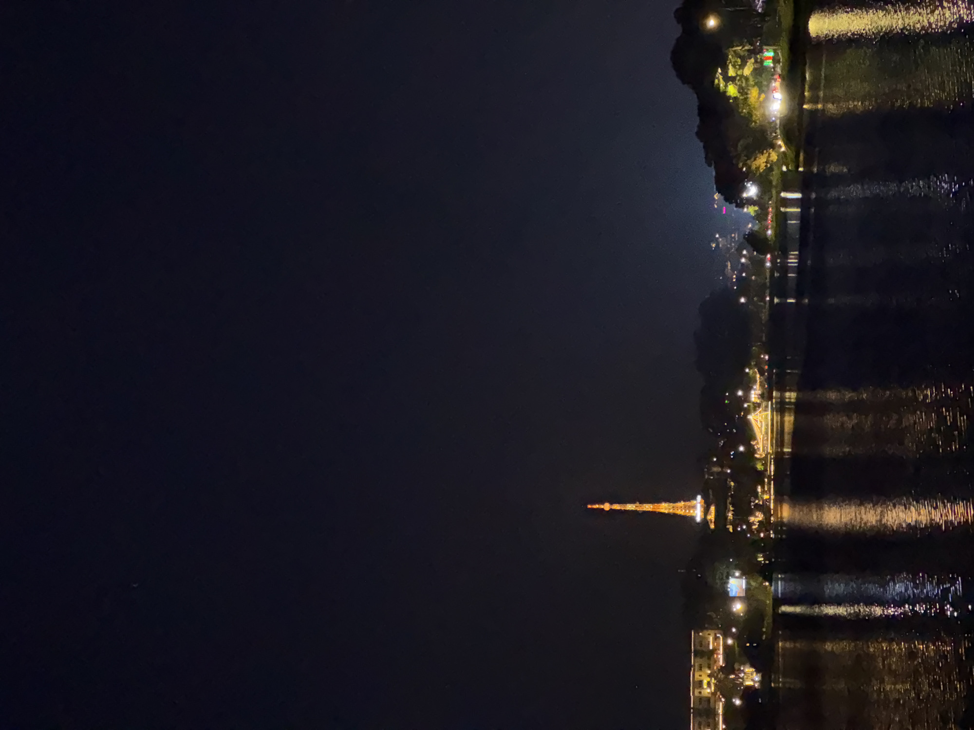At night, a body of water reflects lights from the shoreline and a distant, illuminated tower in Da Lat, Vietnam. The dark water surface is rippled, creating streaks of light that mirror the shoreline. The tower, likely the Da Lat TV Tower, is a slender, lit structure on the left. Along the shoreline, strings of lights delineate the edge, and buildings and trees are visible. The overall scene has a serene, nocturnal atmosphere.