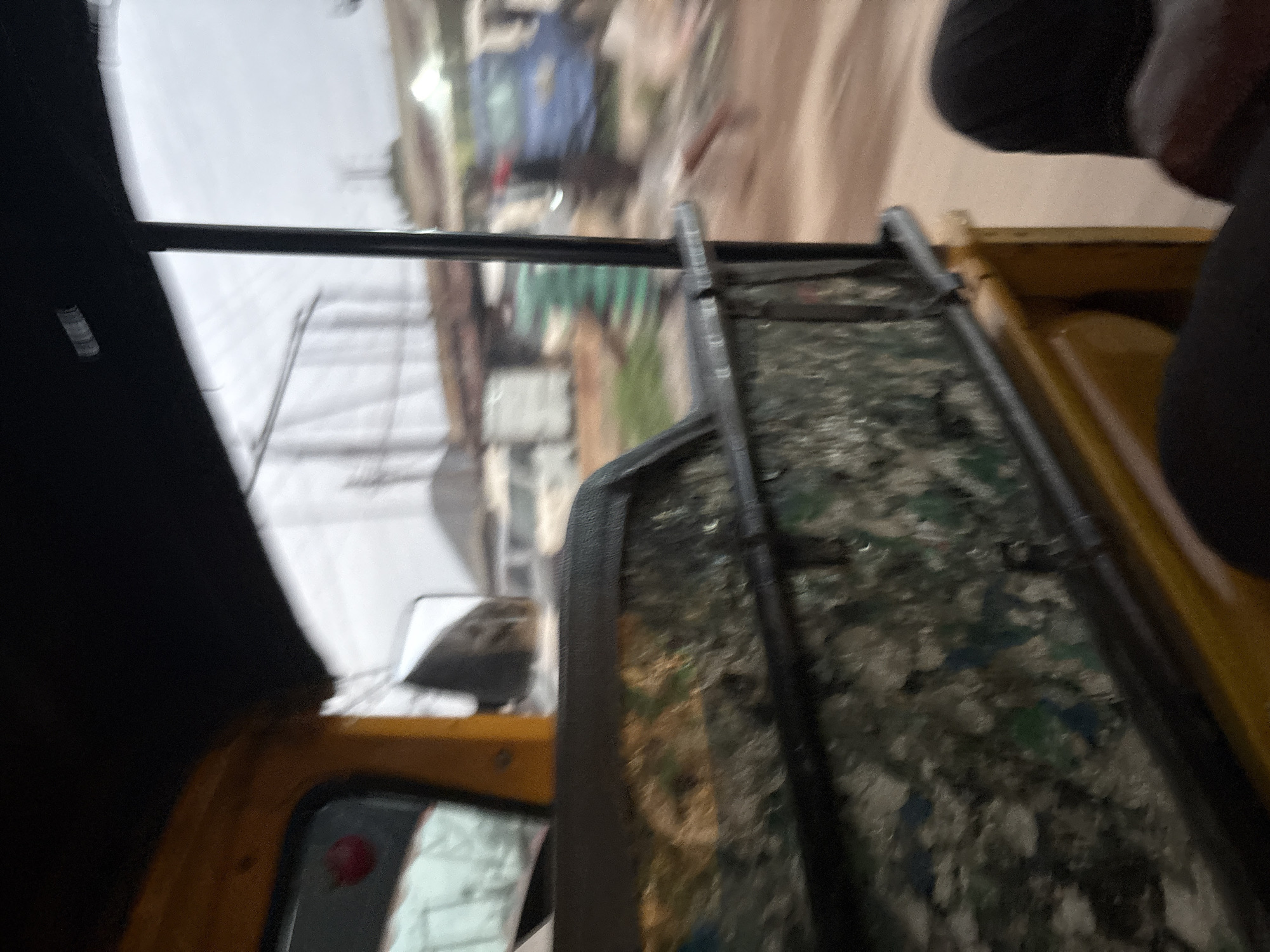 The image captures an interior view from a yellow motorized rickshaw (a type of auto-rickshaw common in Onitsha, Nigeria). The viewpoint is from the passenger seating area, looking forward and to the right. The passenger's legs, clad in dark fabric, are visible in the lower right corner. The rickshaw's interior features a rough, textured surface on the side panels, decorated with a mosaic of what appears to be recycled materials in shades of green, blue, and white, interspersed with darker elements. A black metal bar runs horizontally across the interior, likely a safety feature or part of the structure.
Outside the rickshaw, the environment is blurred due to motion, suggesting the vehicle is in transit. The scene depicts an urban setting in Onitsha, Nigeria. Structures resembling buildings with corrugated metal roofs are visible in the background, along with power lines and utility poles. Several vehicles are present, including a white truck or van and a blue vehicle. A person wearing a green garment is partially visible near the blue vehicle. The ground appears to be unpaved, with a reddish-brown hue, indicative of a dirt road or pathway. The sky is overcast and appears cloudy.