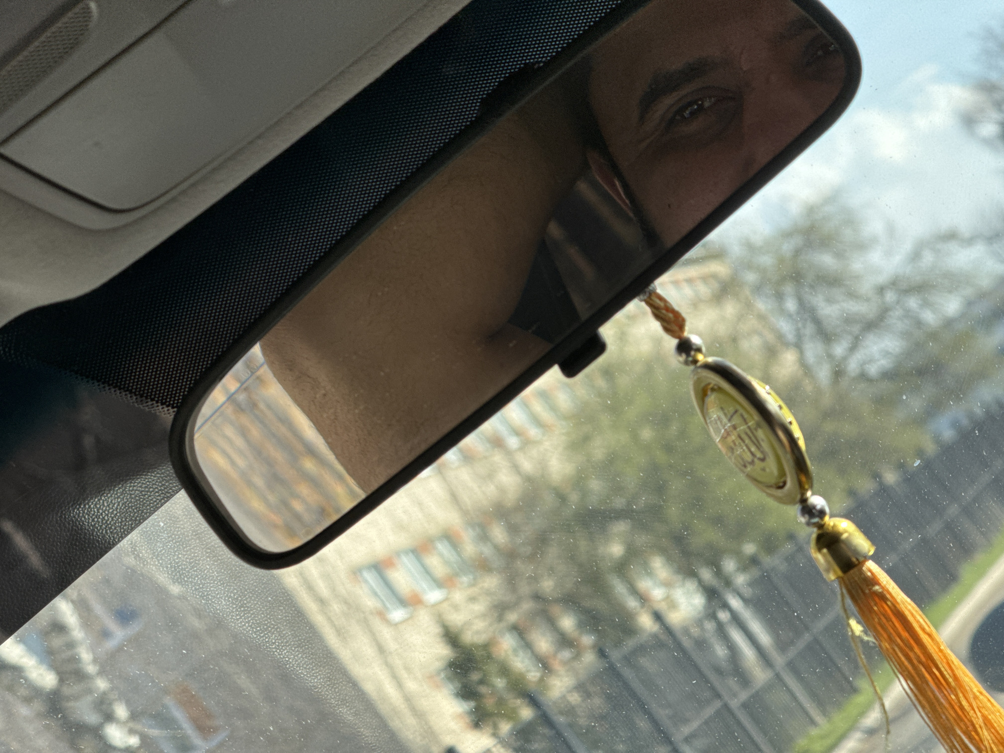 The image captures a perspective from inside a vehicle, looking towards the front windshield and rearview mirror during what appears to be daytime. The primary subject is the rearview mirror, which reflects a portion of a person's face, specifically one eye and part of the forehead, suggesting the driver or a front-seat passenger. The mirror also reflects a blurred view of the car's interior.
Hanging from the rearview mirror is a decorative ornament featuring a golden tassel and a round, metallic pendant. The pendant has intricate designs and what appears to be Arabic calligraphy on its surface, although the specific text is not clearly legible.
Through the windshield, a blurred exterior scene is visible. This includes the light blue sky with some clouds, bare trees, and multi-story buildings, indicating an urban or suburban setting. The windshield itself shows numerous small speckles, suggesting dust or dirt on its surface. The car's headliner and sun visor area are visible above the mirror, along with a textured black trim element beneath the headliner. The overall lighting is bright, consistent with daylight. The scene takes place in Chaikovsky, Russia.