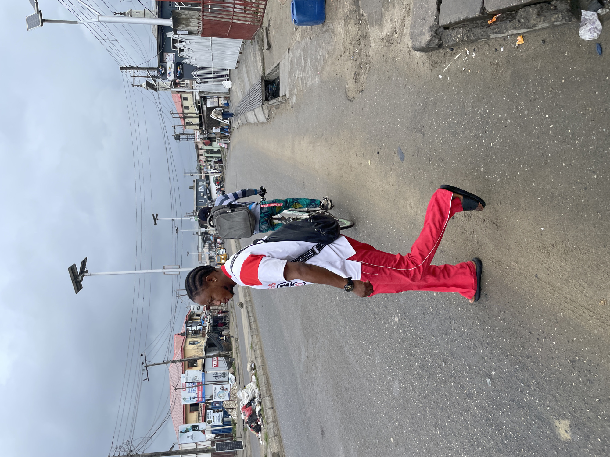 In the city of Ado, Nigeria, under an overcast sky, a person walks across a paved street. The individual is wearing a white t-shirt with red accents on the sleeves and collar, red pants with white stripes down the sides, and black slides. They have a black bag slung across their body. In the background, another person is riding a bicycle, also carrying a backpack. The street is lined with buildings, some appearing to be shops or businesses, and there are power lines and streetlights overhead. Piles of debris are visible on the side of the road. The overall scene depicts a busy urban environment.