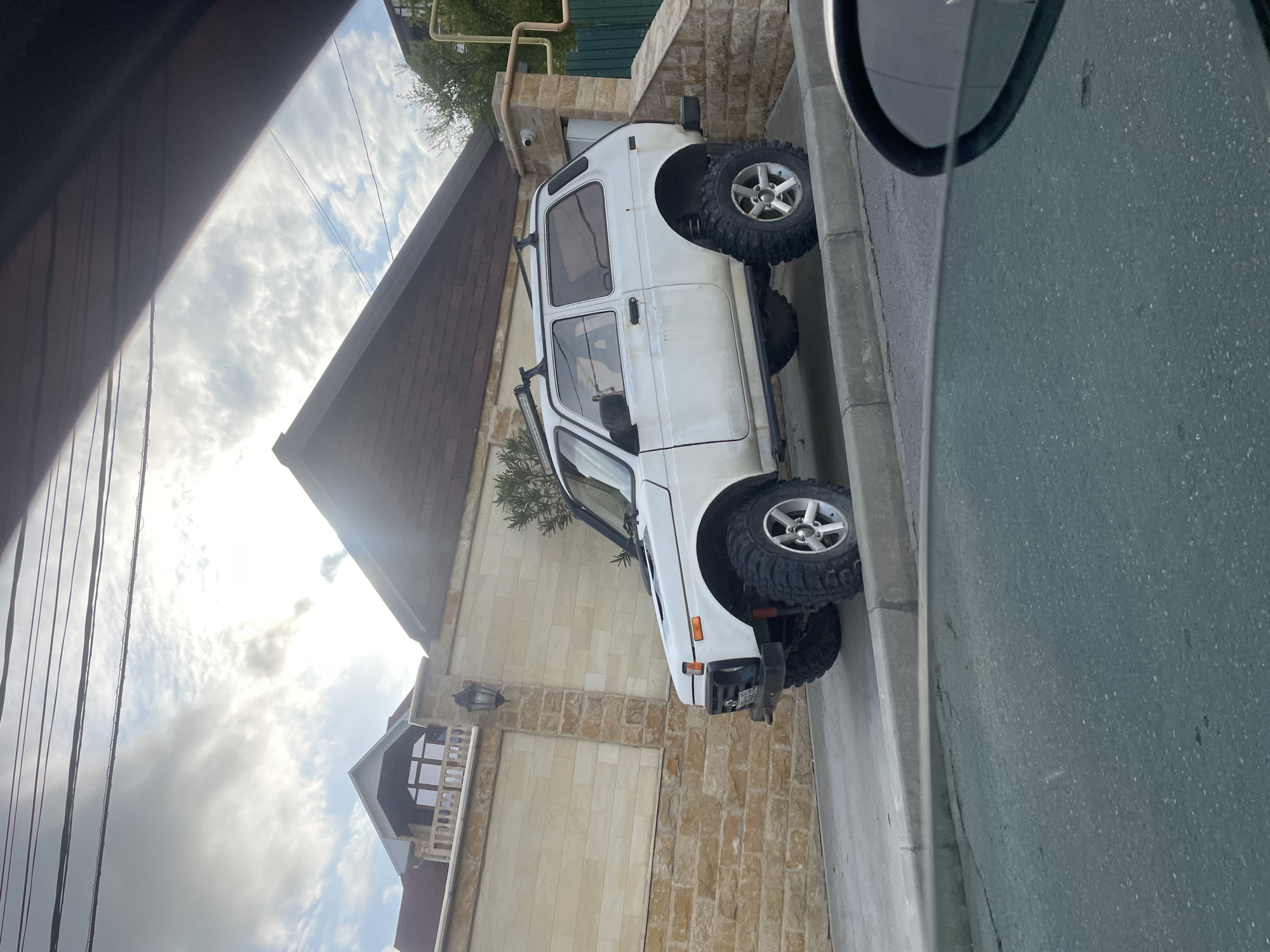 A white, boxy, four-wheel-drive vehicle is parked on the side of a street in what appears to be Baranovka, Russia, based on the style of architecture visible. The vehicle is facing slightly away from the camera. The street is paved and has a curb. A building with a light-colored brick facade stands beside the vehicle. The sky is cloudy. Part of a vehicle with a side mirror is visible in the right foreground, suggesting the photo was taken from inside a car. Power lines are visible in the sky. A security camera is mounted on the wall of the building next to the parked car.