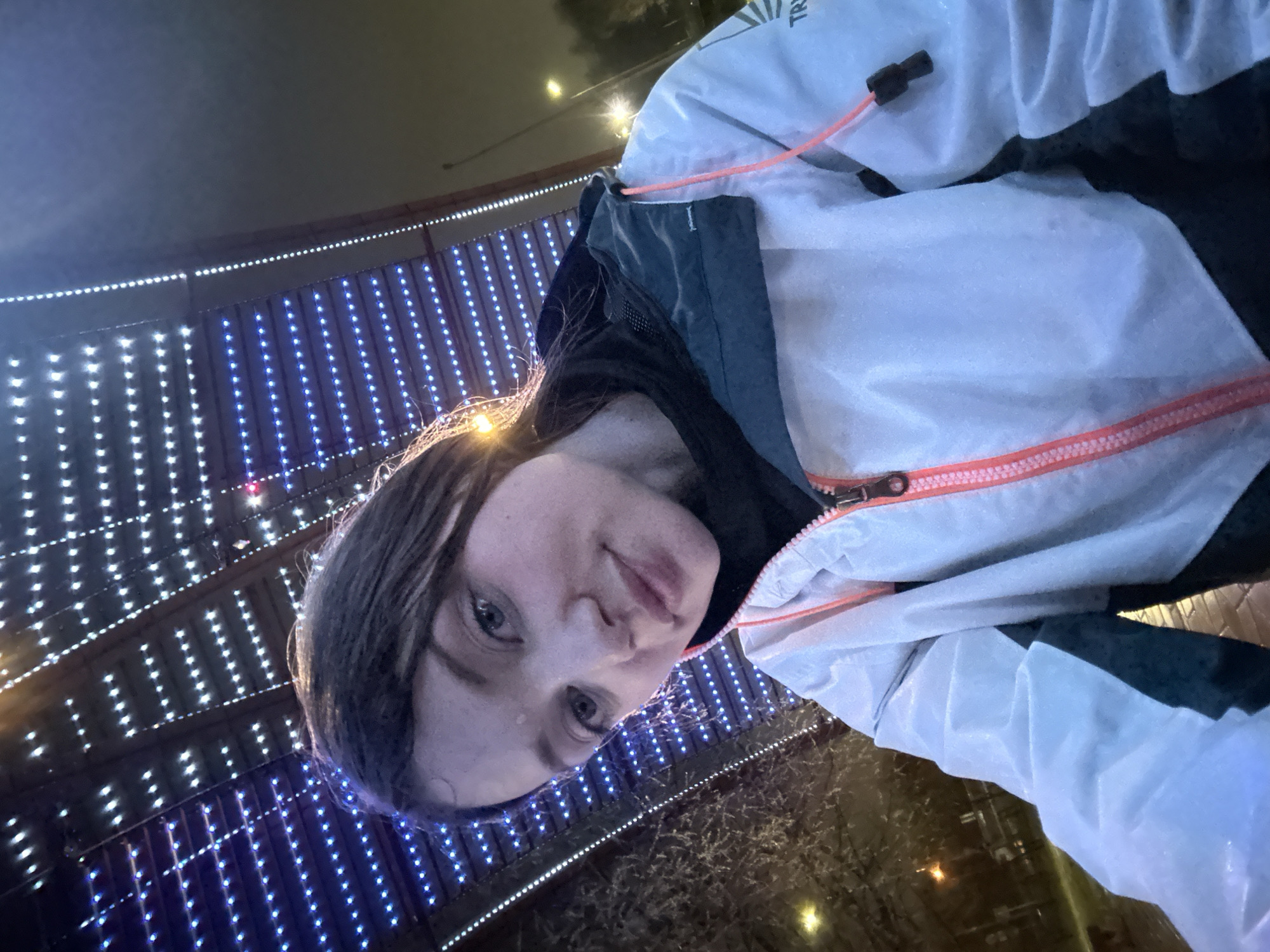 This vertical selfie captures a person outdoors at night, likely in Kazan, Russia. The central subject is a person, presumably a woman, with long dark hair, looking directly at the camera with a neutral expression. Her face and hair appear slightly damp, possibly from mist or light precipitation, with a visible glint of moisture on her cheek.
She is wearing a light-colored, possibly white or very light grey, jacket with a distinct dark blue or grey inner layer. The jacket features vibrant orange details, including a bright zipper and a drawstring with a black toggle. Partially visible on the jacket's upper right shoulder is the text "TR".
The background is a prominent feature, illuminated by numerous strings of LED lights. These lights are arranged in both horizontal and vertical lines, with some displaying cool white light and others emitting a blue hue, creating a striking visual pattern against the dark night sky. The arrangement suggests an illuminated architectural structure, a bridge, or a festive display. In the lower part of the image, hints of dark, possibly wet ground or a reflective surface are visible, alongside sparse, light-colored foliage that could be snow-dusted or frosted, reinforcing the impression of a cold or damp evening. The bright artificial lighting strongly indicates the time of day is night.