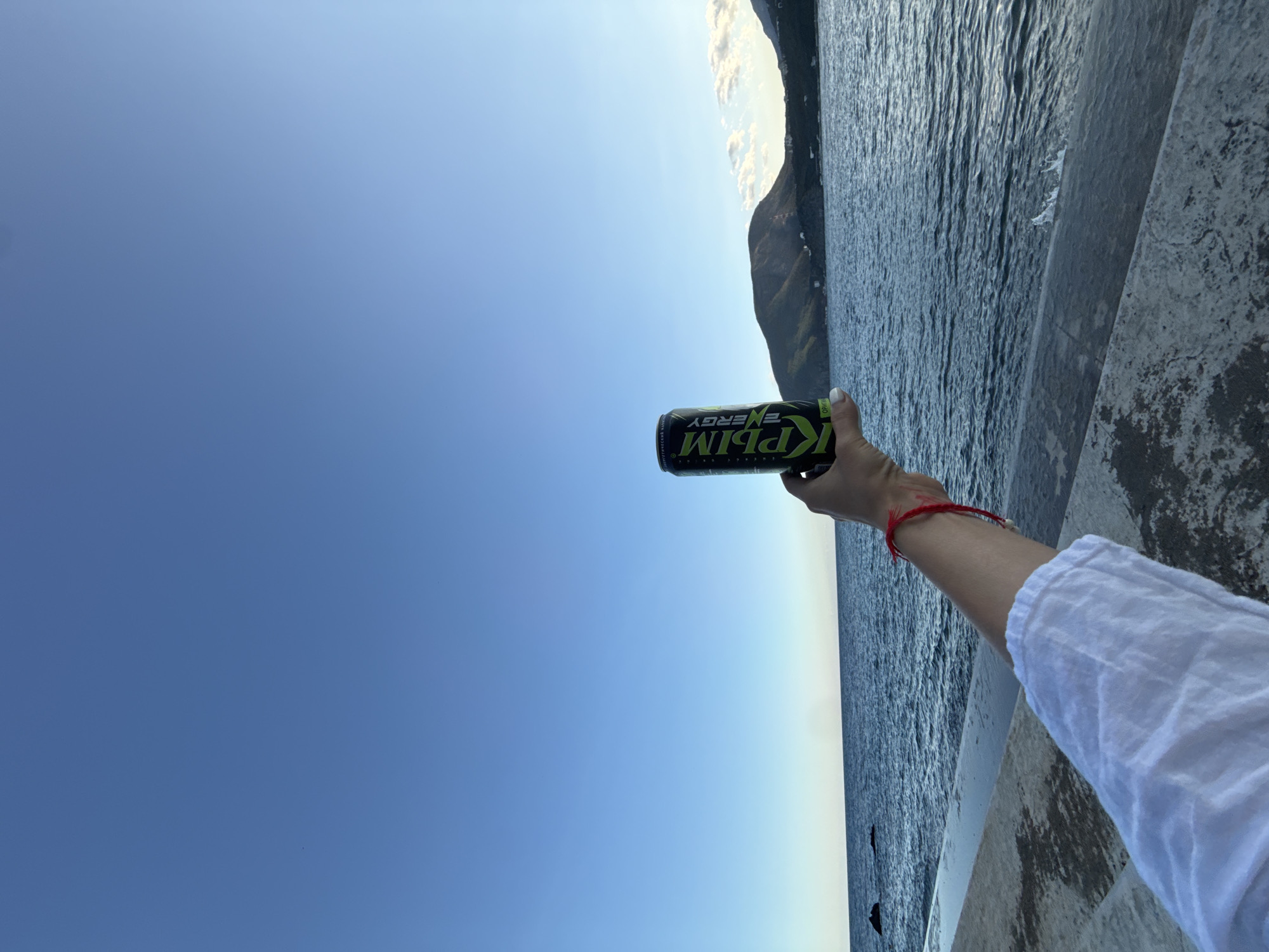 An individual's left arm, adorned with a red string bracelet, holds a black and green can of "КРЫМ ENERGY" above a body of water. The can is positioned in the foreground, slightly right of center. Behind the can, a vast expanse of blue water stretches towards a distant coastline with rolling hills under a clear, bright blue sky. The water appears calm with gentle ripples. The foreground shows a concrete walkway or embankment. The scene suggests an outdoor setting, possibly a waterfront promenade.