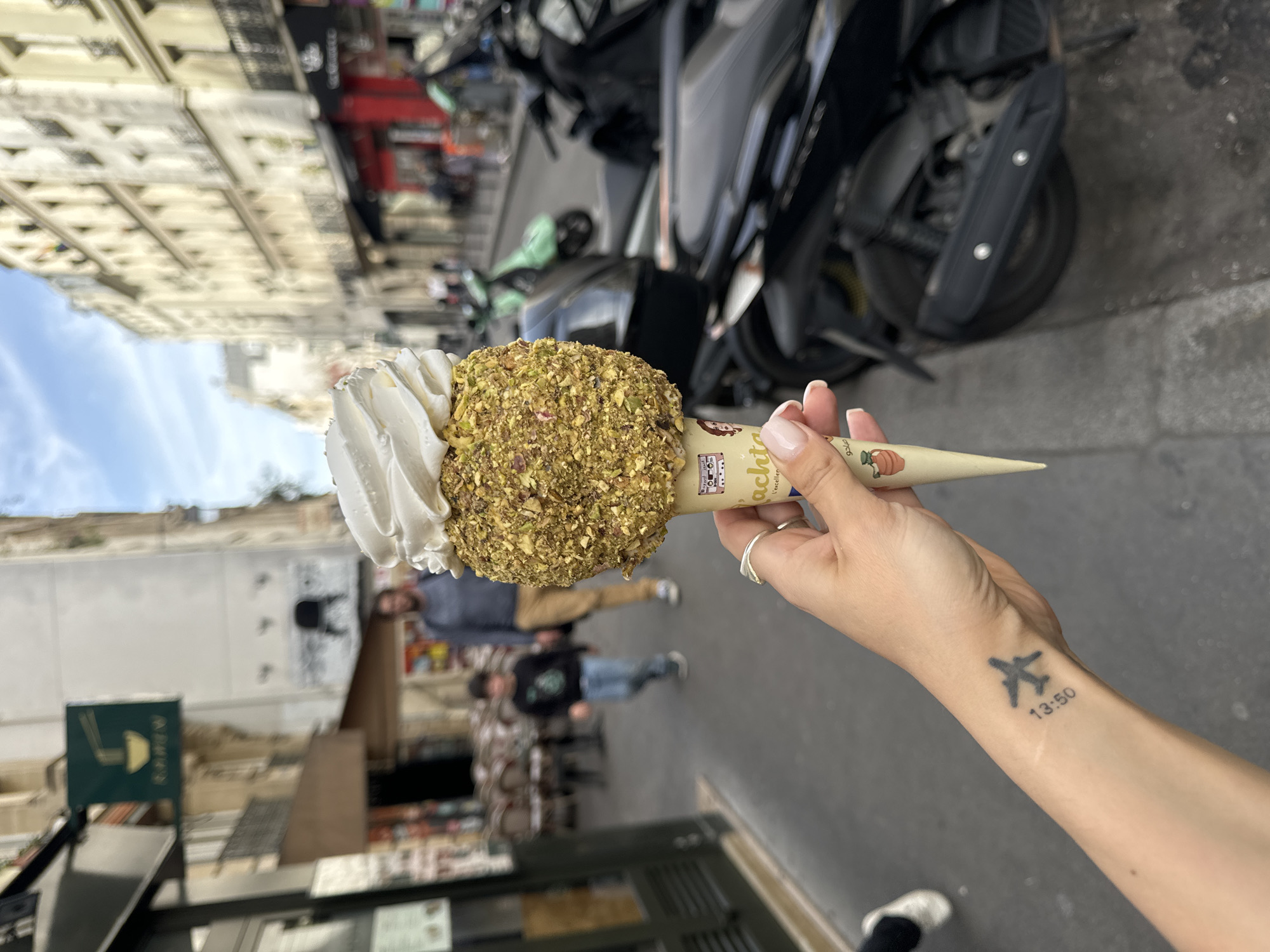 A medium shot, taken at eye-level, shows a person's hand holding an ice cream cone in the city of Paris, France. The ice cream has two scoops. One scoop is white with a swirl, and the other is coated in small green and brown nuts. The cone is beige with illustrations. The person's hand has a silver ring and manicured nails. An airplane tattoo with the numbers "13:50" is visible on their left forearm. The background is a blurred Parisian street scene with pedestrians, parked motorcycles, and buildings.
