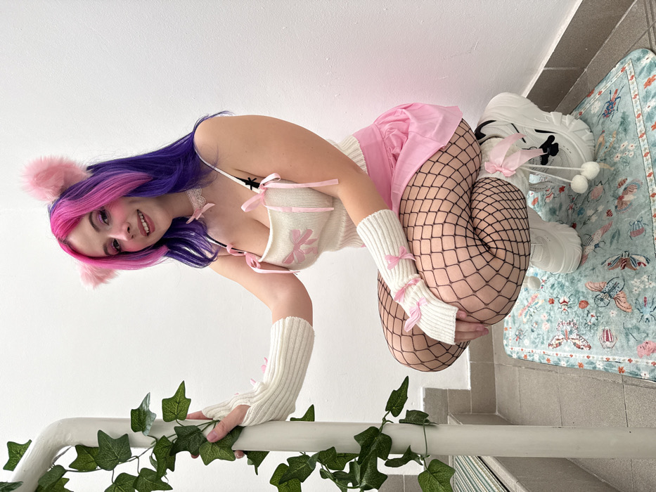 The image displays a young person posed in what appears to be a playful and artistic style, rotated 90 degrees clockwise.
The central figure is a person with light skin, wearing a vibrant and eclectic outfit. Their long hair is dyed in two prominent colors: a bright, hot pink on the front and top layers, blending into a deep purple for the majority of the hair. They are wearing a pink, fluffy cat-ear headband. Their face is turned towards the camera, and they are smiling with their lips slightly parted. Visible makeup includes what appears to be pink blush or eyeshadow around the eyes.
Their attire consists of several distinct pieces:
* A cream or off-white knitted crop top or bralette with thin spaghetti straps. It features intricate detailing including pink ribbon ties on the shoulders and across the front, forming small bows. A delicate pink flower design is embroidered or appliquéd on the chest.
* A delicate white lace choker or necklace is worn around their neck.
* A short, light pink pleated skirt is worn over black fishnet stockings that cover both legs.
* They wear cream or off-white knitted fingerless arm warmers on both arms, each adorned with a small pink bow near the wrist.
* Their footwear consists of chunky white sneakers with thick soles. One visible shoe has black design elements and white shoelaces that are decorated with small, white fluffy pom-poms.
The person is in a dynamic, crouching or kneeling pose. Their left hand (from the viewer's perspective) is gently gripping a white railing or banister. This railing is decorated with artificial green ivy, creating a natural accent. Their right leg is bent and drawn up, with the fishnet-clad knee visible.
The setting appears to be indoors, possibly on a staircase or landing. In the background, there is a plain white wall. The floor beneath them is a light grey tile. On the floor, a rectangular rug with a light blue-green background is visible, patterned with intricate designs of butterflies, leaves, and floral motifs in shades of pink, orange, and blue.
The overall impression is whimsical and "kawaii," characterized by the pastel colors, animal ears, and playful accessories. The lighting is bright and even, highlighting the details of the outfit and the person's expression. No text is visible in the image.